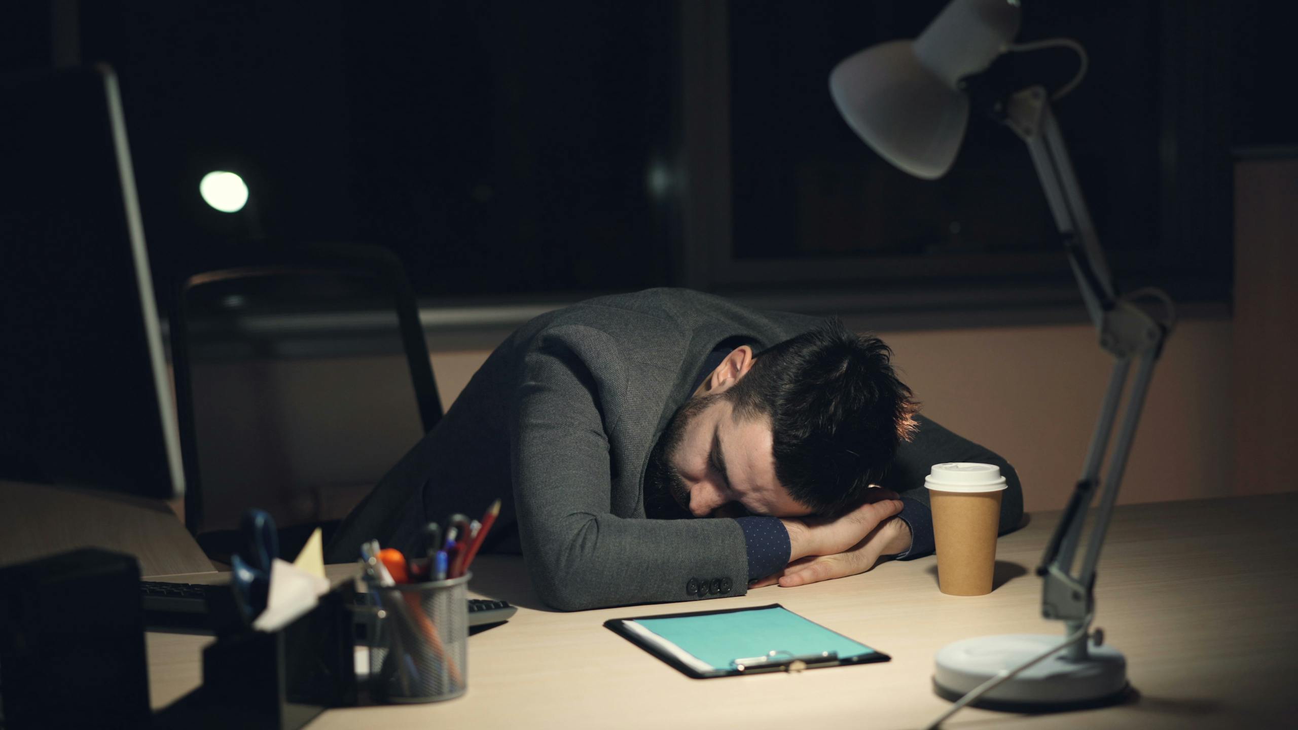 Man in office suit sleeping on desk with coffee and tablet nearby, symbolizing work fatigue.