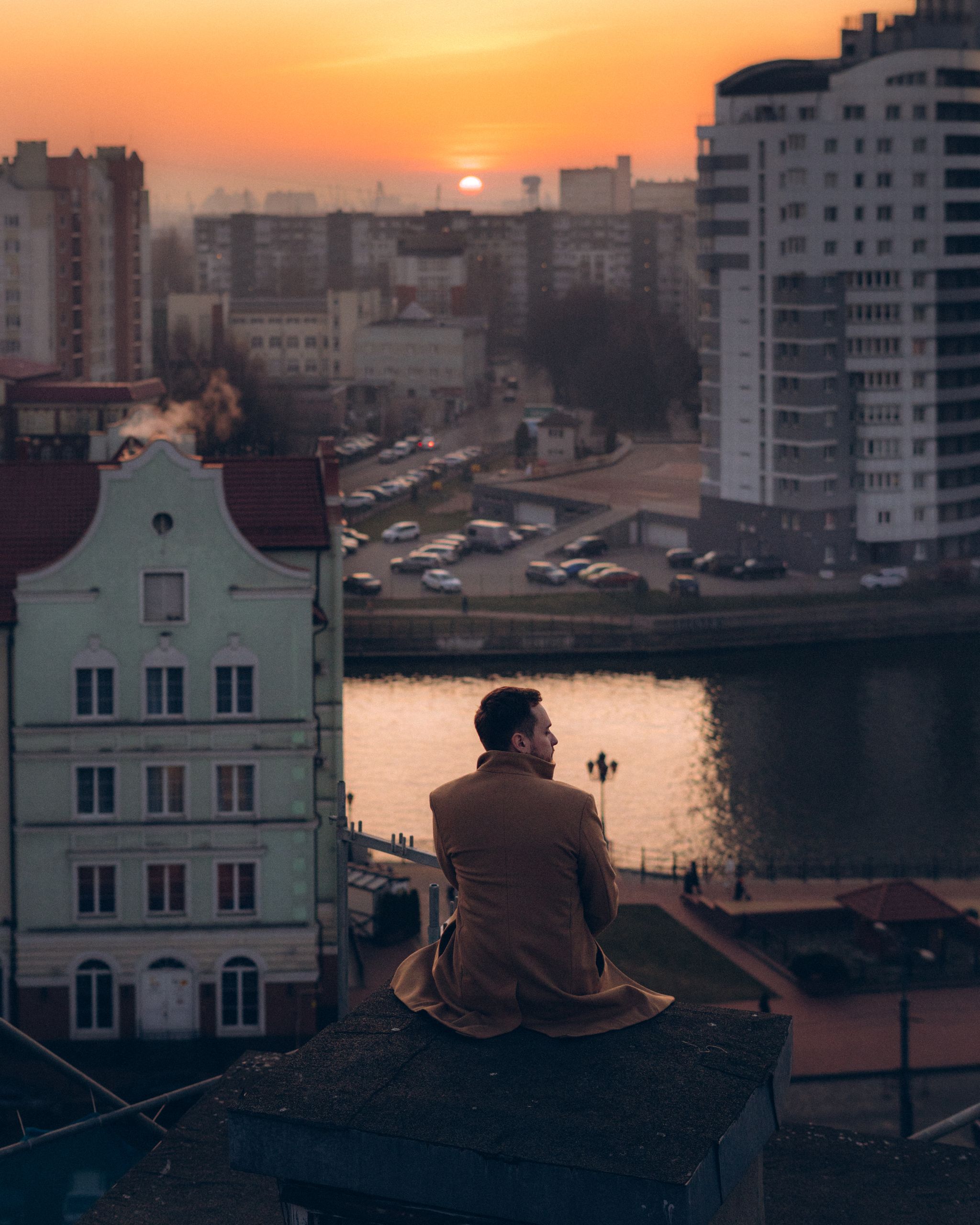 Man in warm coat sitting on rooftop, contemplating a vibrant sunset over a cityscape.