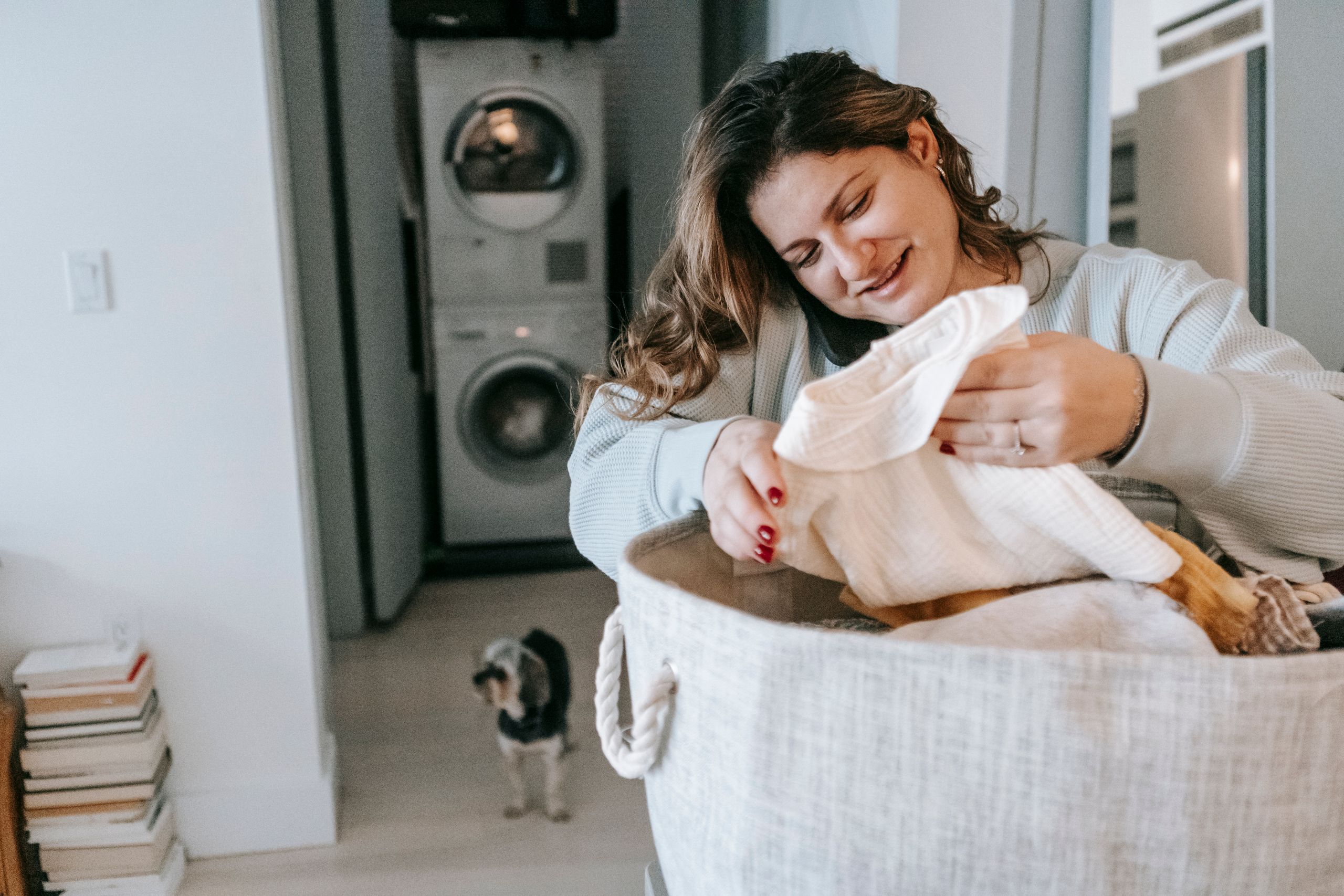 Optimistic female with clean clothes in basket having phone conversation while standing in light washroom with washing machines and dog