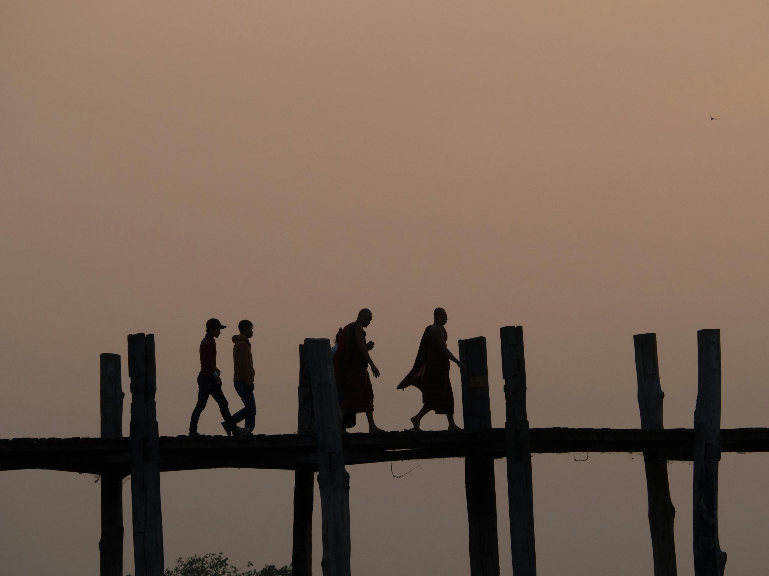 People crossing U Bein Bridge in Mandalay, Myanmar, during dusk, creating serene silhouettes against the sunset.