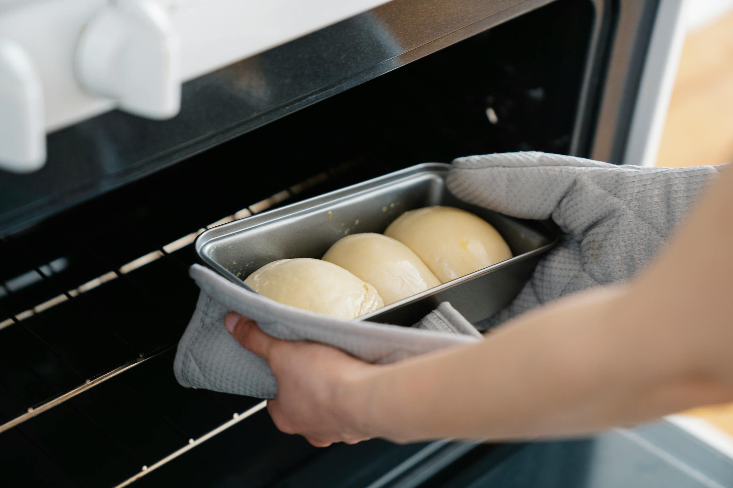 Person placing dough buns in oven, wearing gloves and using baking tray. Captured mid-preparation in a kitchen setting.