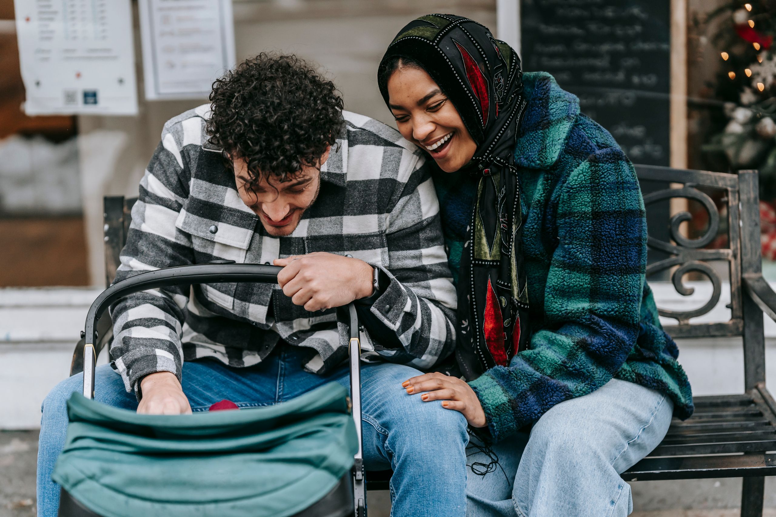 Playful ethnic couple sitting close on bench and having fun with baby lying in stroller