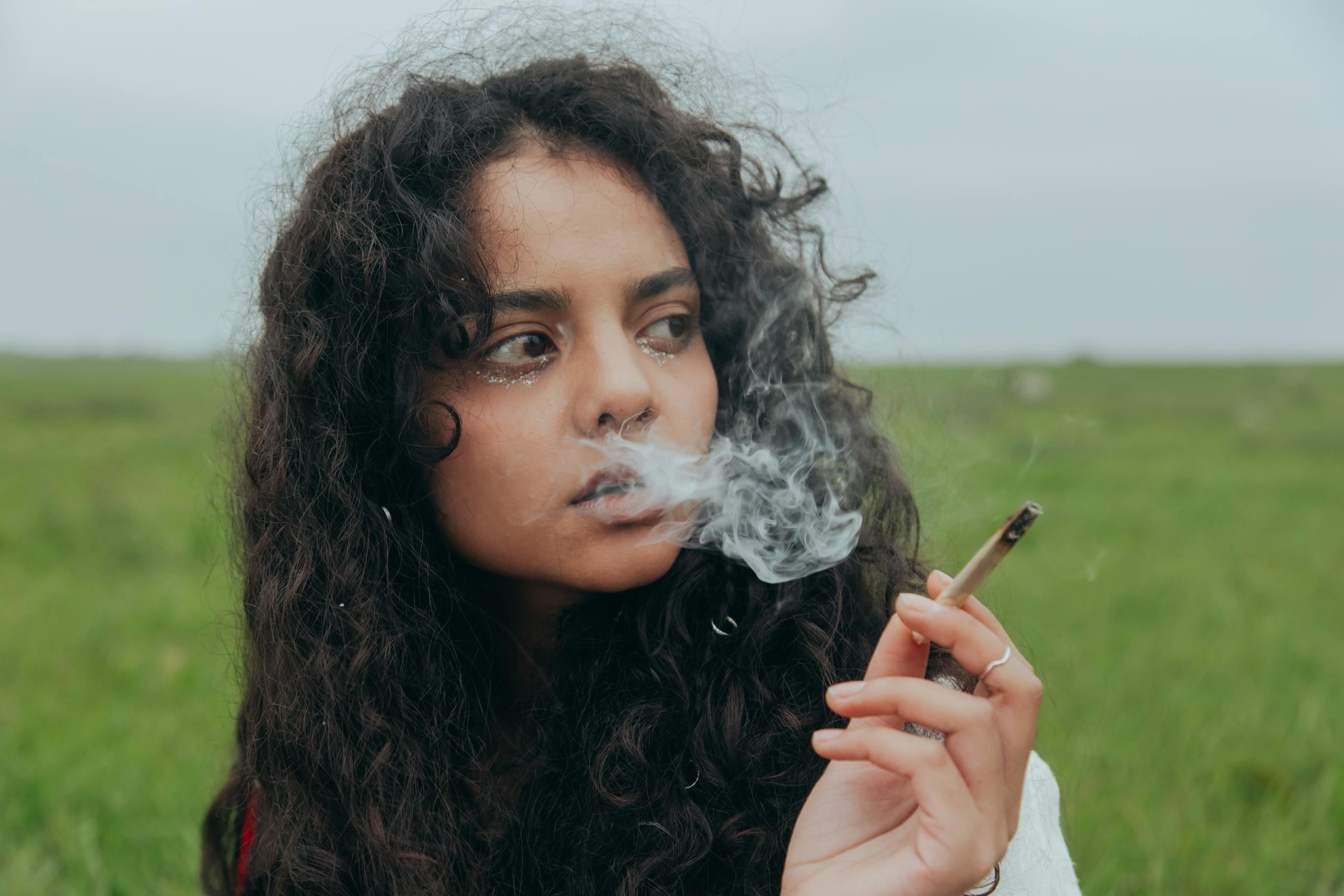 Portrait of a young woman with curly hair smoking in a grassy field, evoking a pensive mood.