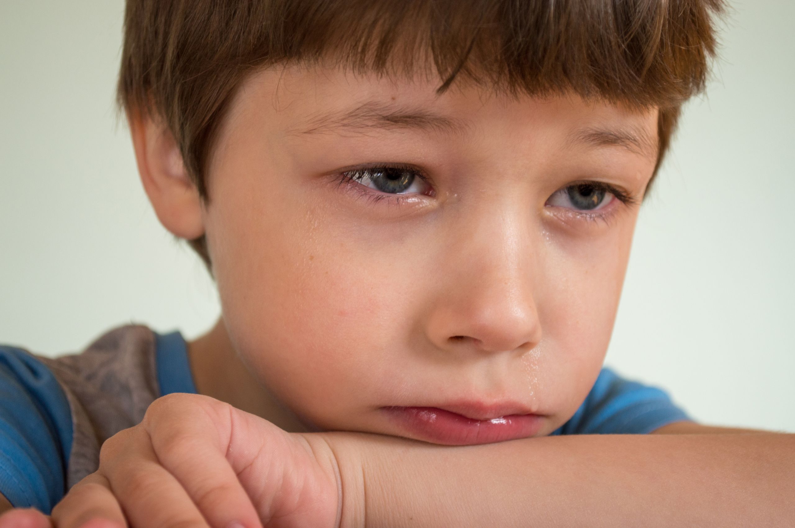 Portrait of crop upset little boy with brown hair and green eyes crying and resting head on arm while looking away against white background