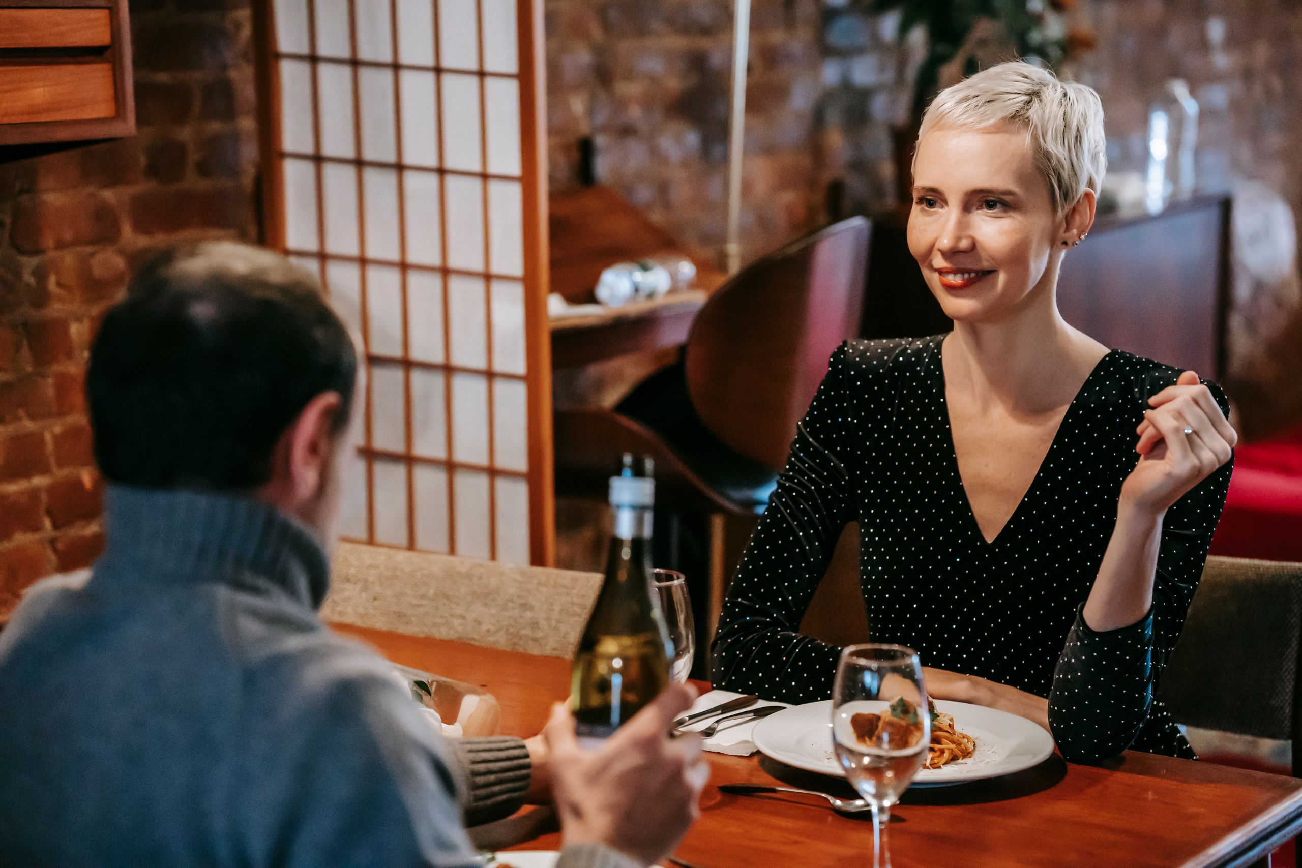 Positive couple sitting at table while having date in bright restaurant with pasta with meatballs on plate with glasses and wine bottle near chairs