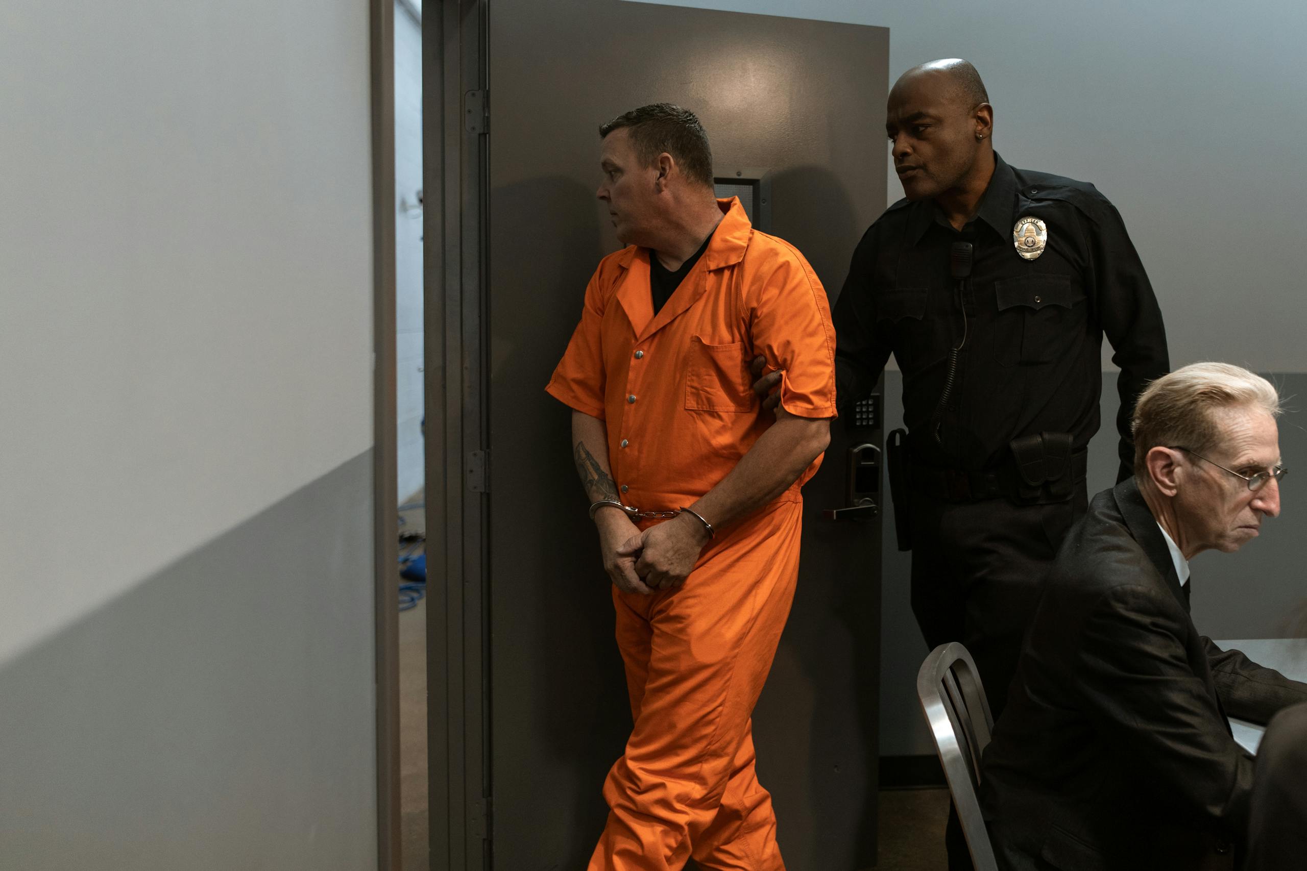 Prisoner in orange jumpsuit being escorted by a correctional officer through facility.