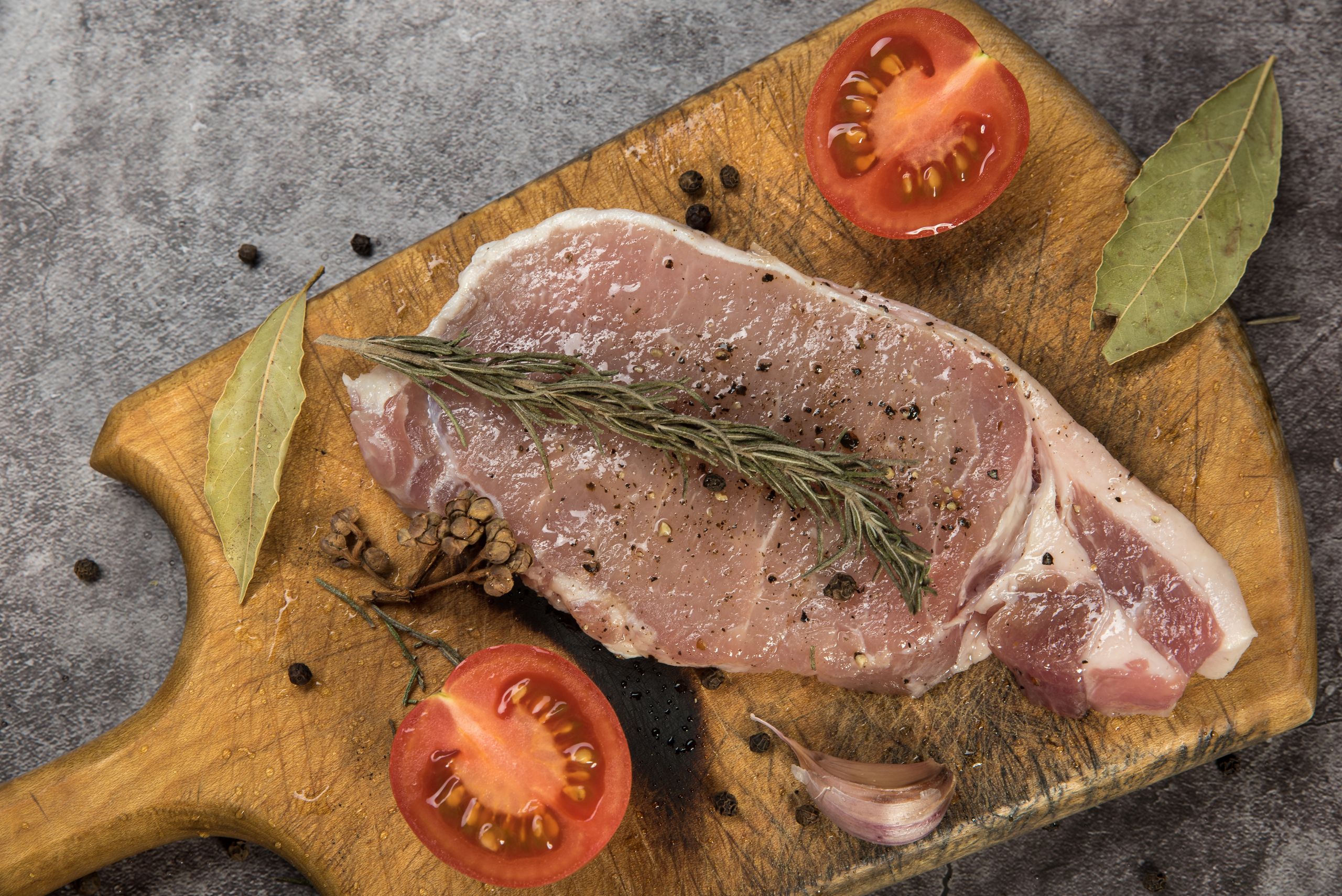 Raw pork chop with seasoning, herbs, and tomatoes on a wooden board.