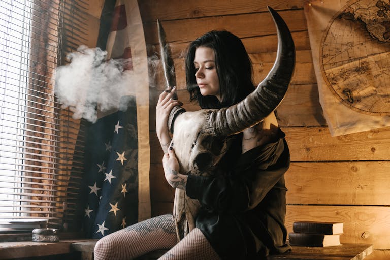 Relaxed brunette with tattoos and nose piercing in short skirt smoking cigarette and breathing out cloud of smoke while sitting on desk and embracing horned animal skull