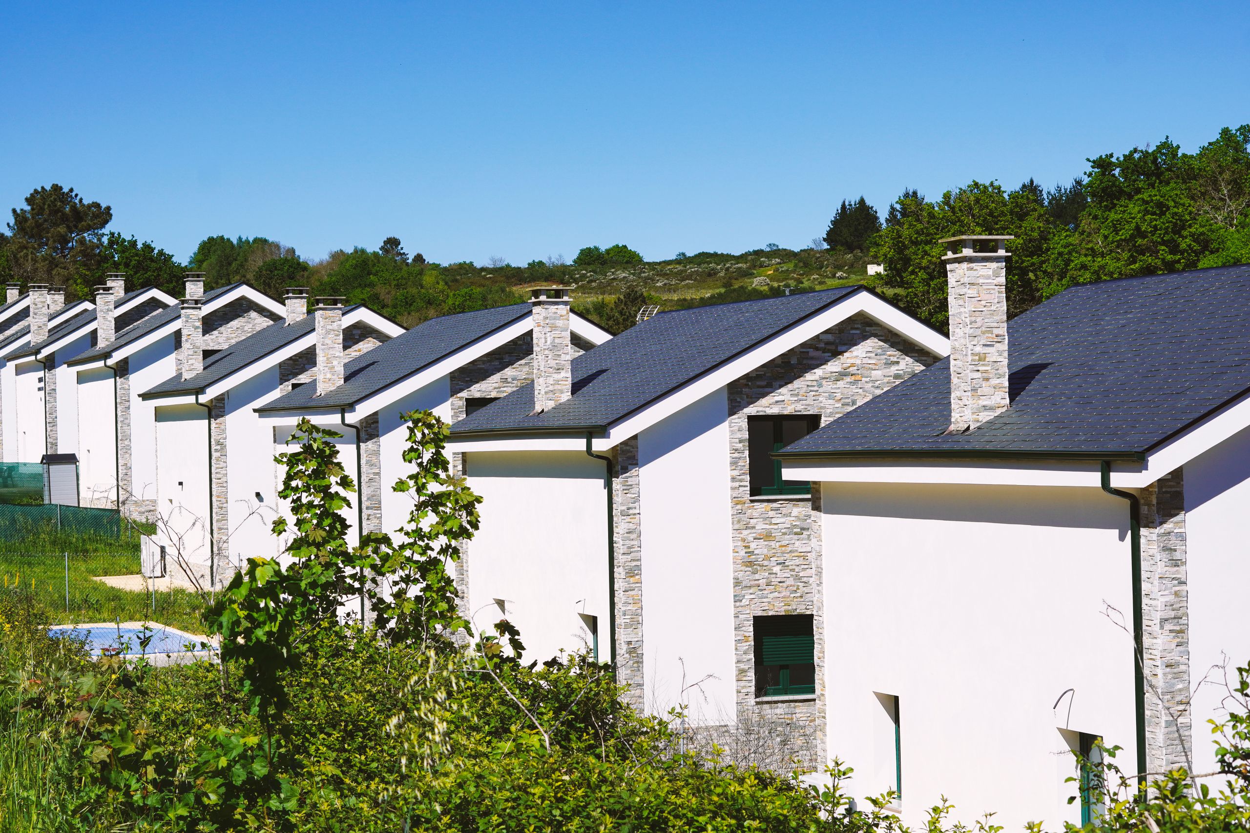 Row of modern suburban houses with chimneys in a sunny rural setting, surrounded by greenery.