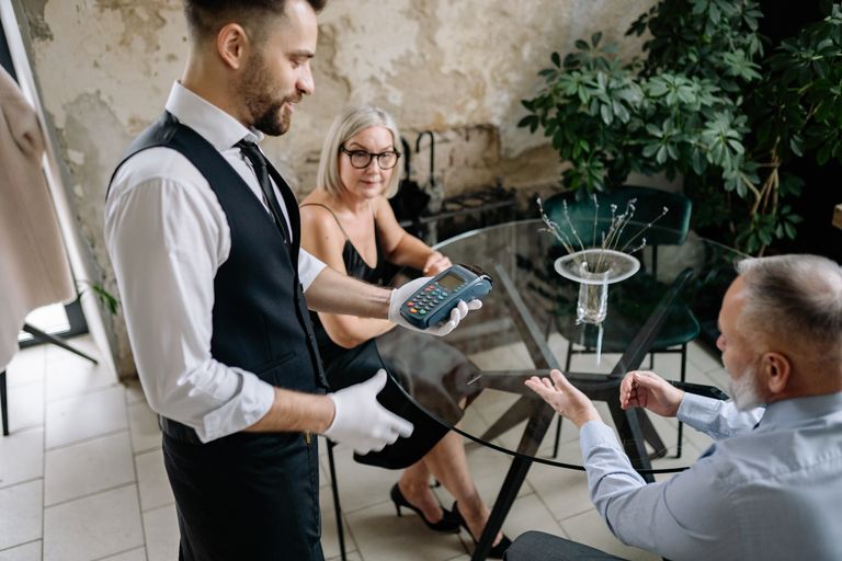 Senior couple completes payment with waiter in stylish restaurant setting, showcasing hospitality service.