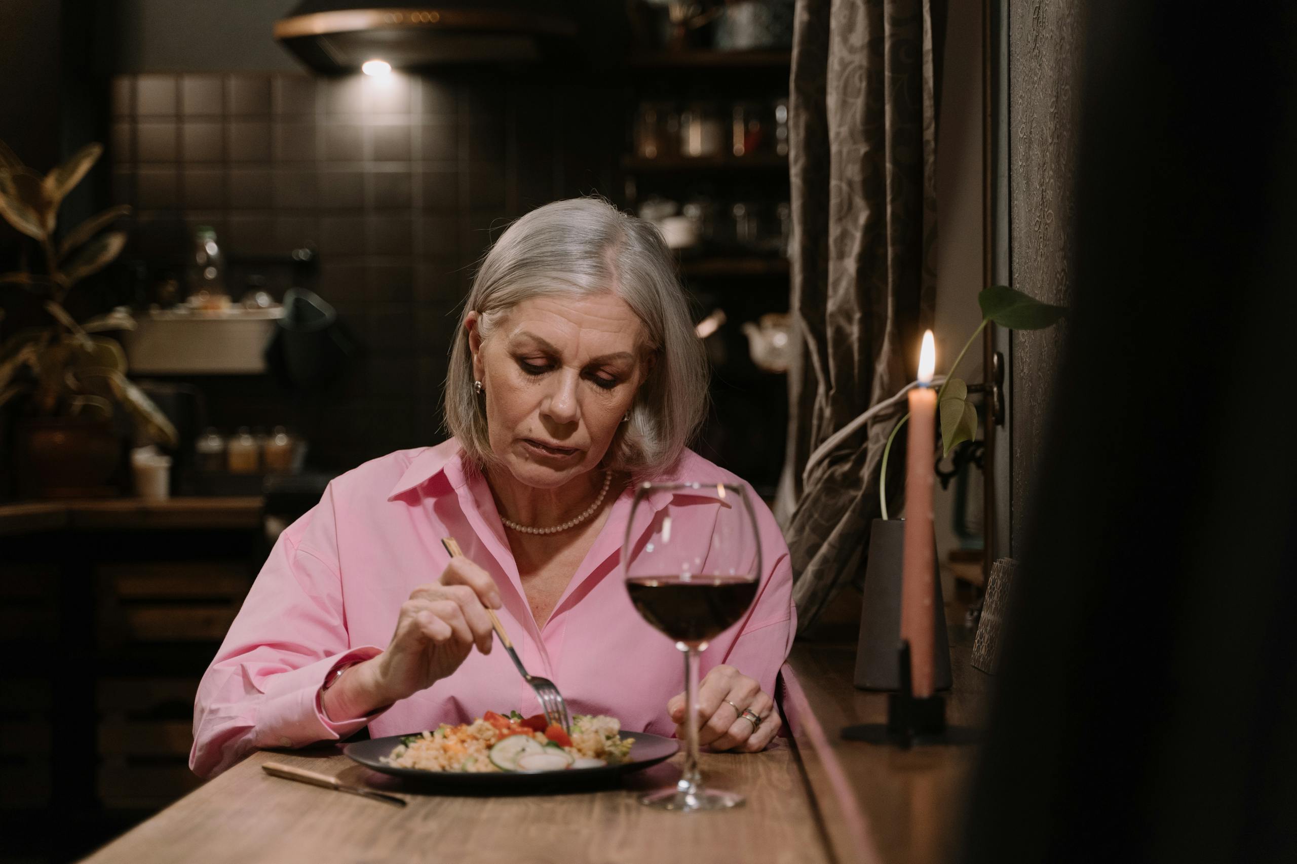 Senior woman enjoys a solitary candlelit dinner with red wine in a cozy indoor setting.