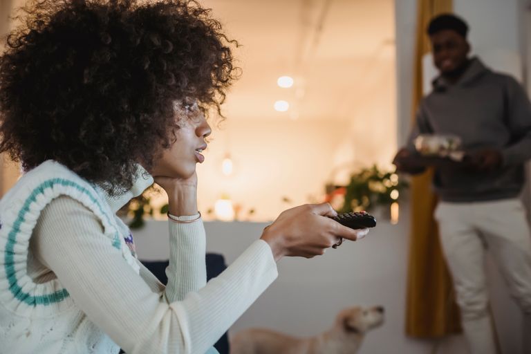Side view of attentive young African American lady with curly hair switching channels on TV with remote controller while sitting on sofa with hand at chin near smiling boyfriend