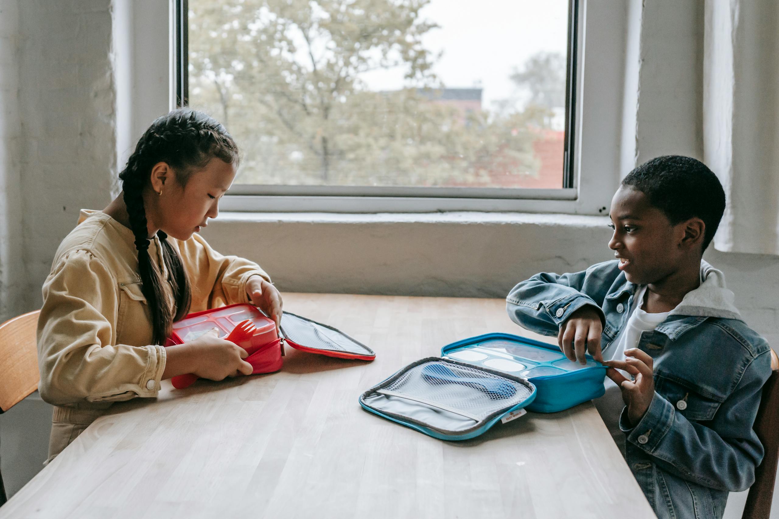 Side view of diverse children in casual clothes sitting at table with lunch boxes during break in school