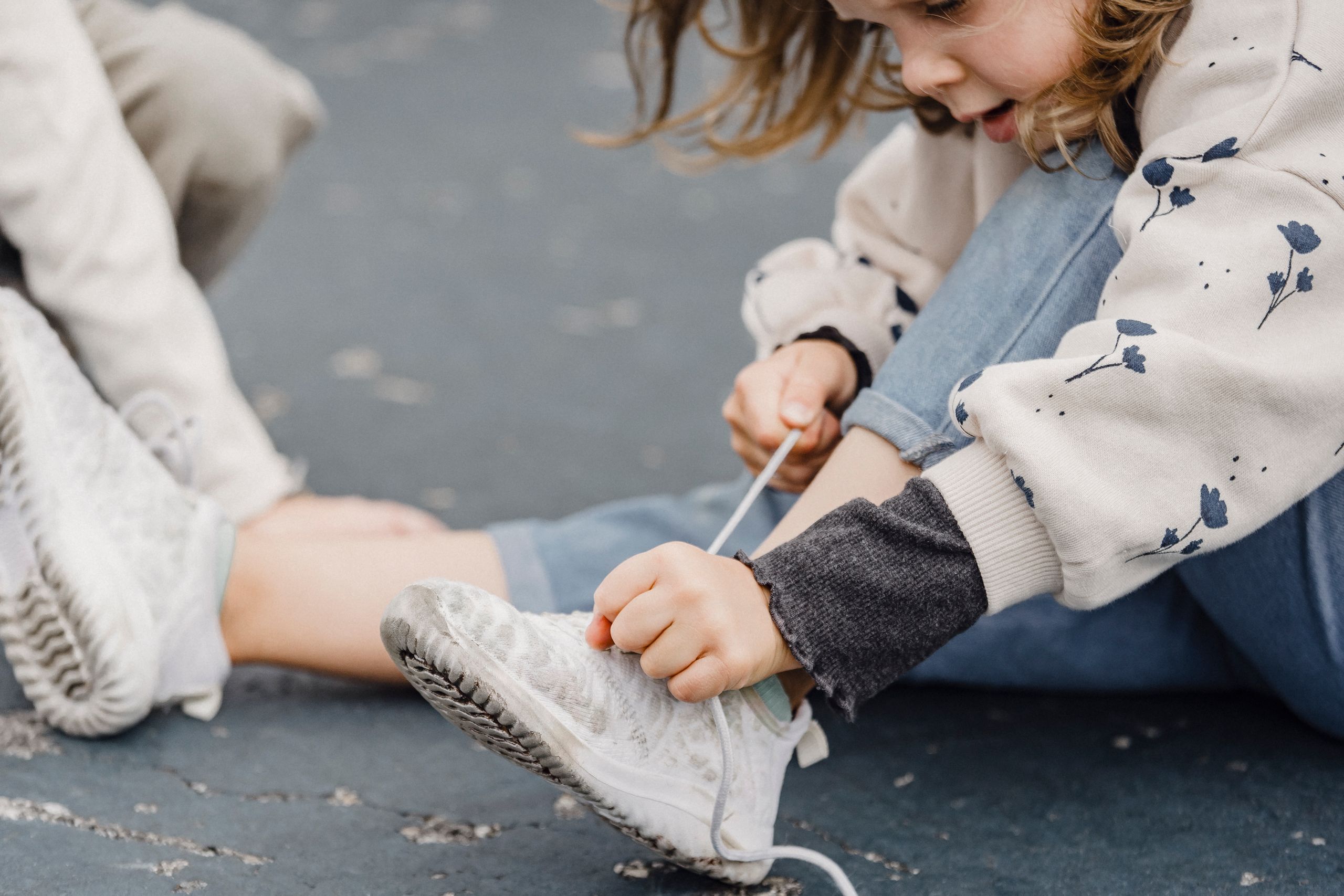 Side view of focused girl tying shoelaces on footwear while sitting with bent leg on playground near crop kid on blurred background
