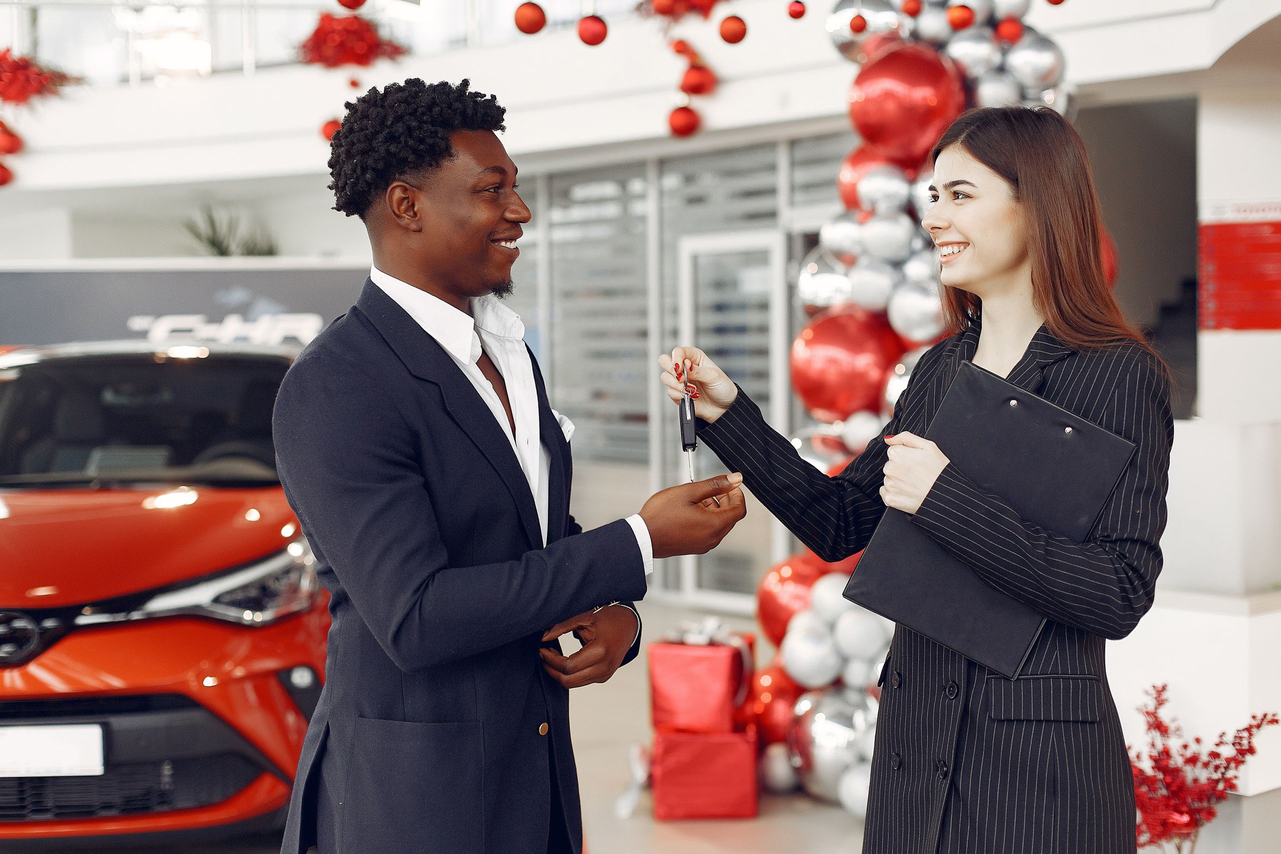 Side view of happy positive female dealer in formal classy wear passing car keys to excited African American customer in trendy suit in car showroom