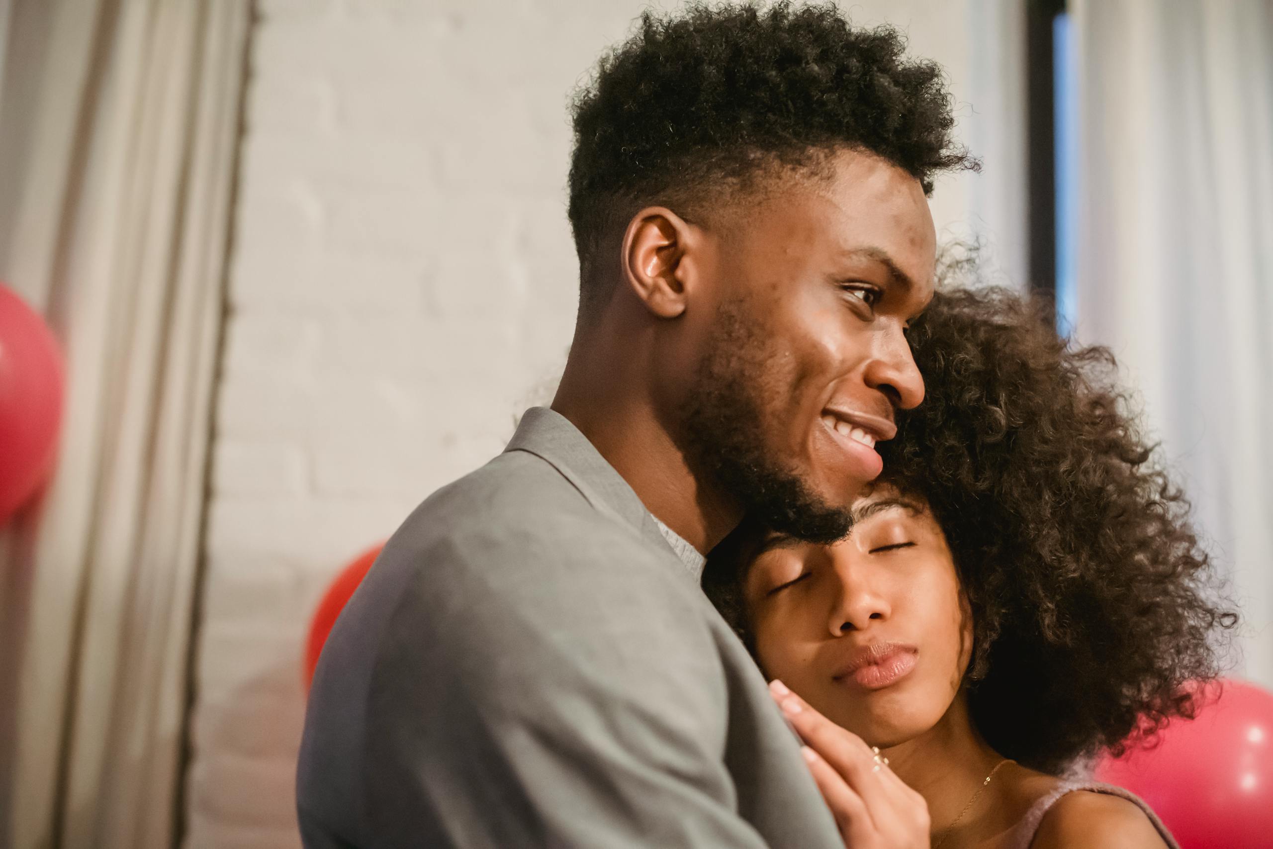 Side view of young African American couple in elegant outfits embracing and dancing while standing and leaning on each other in bright apartment near balloons on wall and curtains