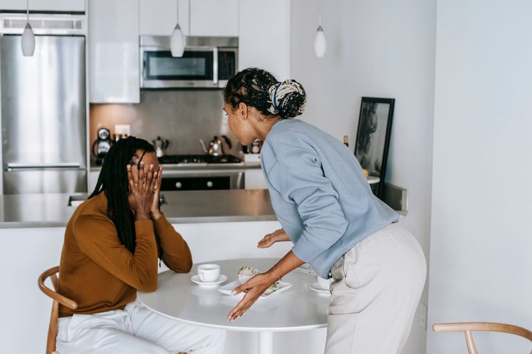 Side view of young black guy sitting at table with hands on head while arguing with standing near table lady at home
