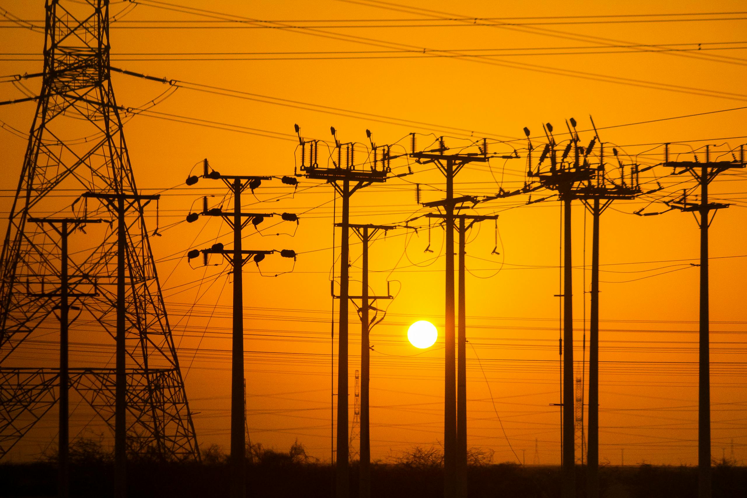 Silhouette of electric towers during sunset, highlighting power lines with a warm orange sky.