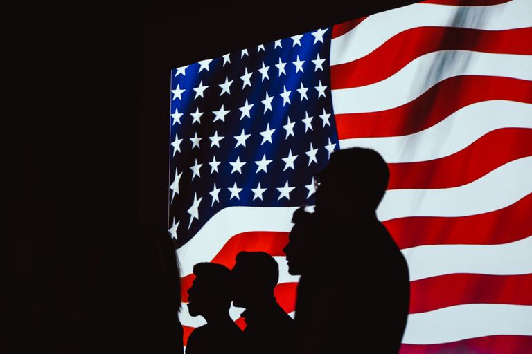 Silhouetted figures in front of a bright, wavy American flag create a patriotic scene.