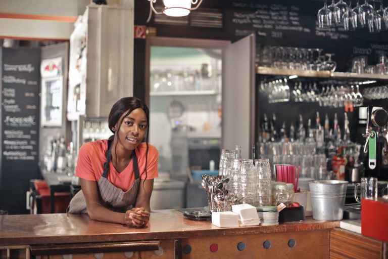 Smiling bartender wearing an apron at a cozy café counter with glasses and utensils.