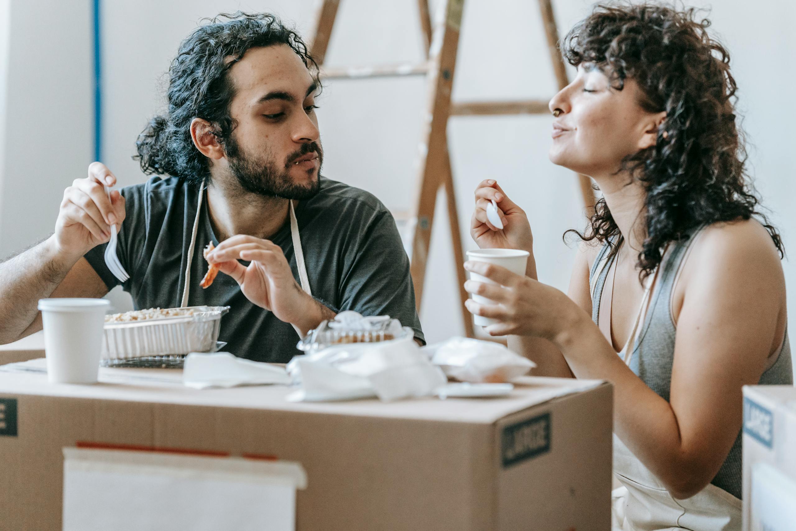 Smiling ethnic woman with closed eyes and coffee against boyfriend with tasty food to go on cardboard box at home