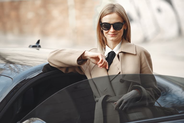 Smiling female driver in stylish coat and sunglasses leaning on automobile while standing on city street with paper bags