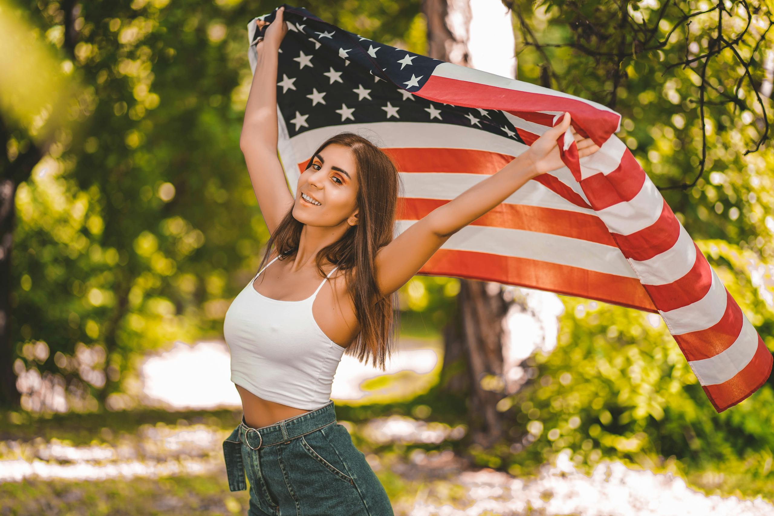Smiling woman in casual attire posing happily with an American flag in a lush green outdoor setting.