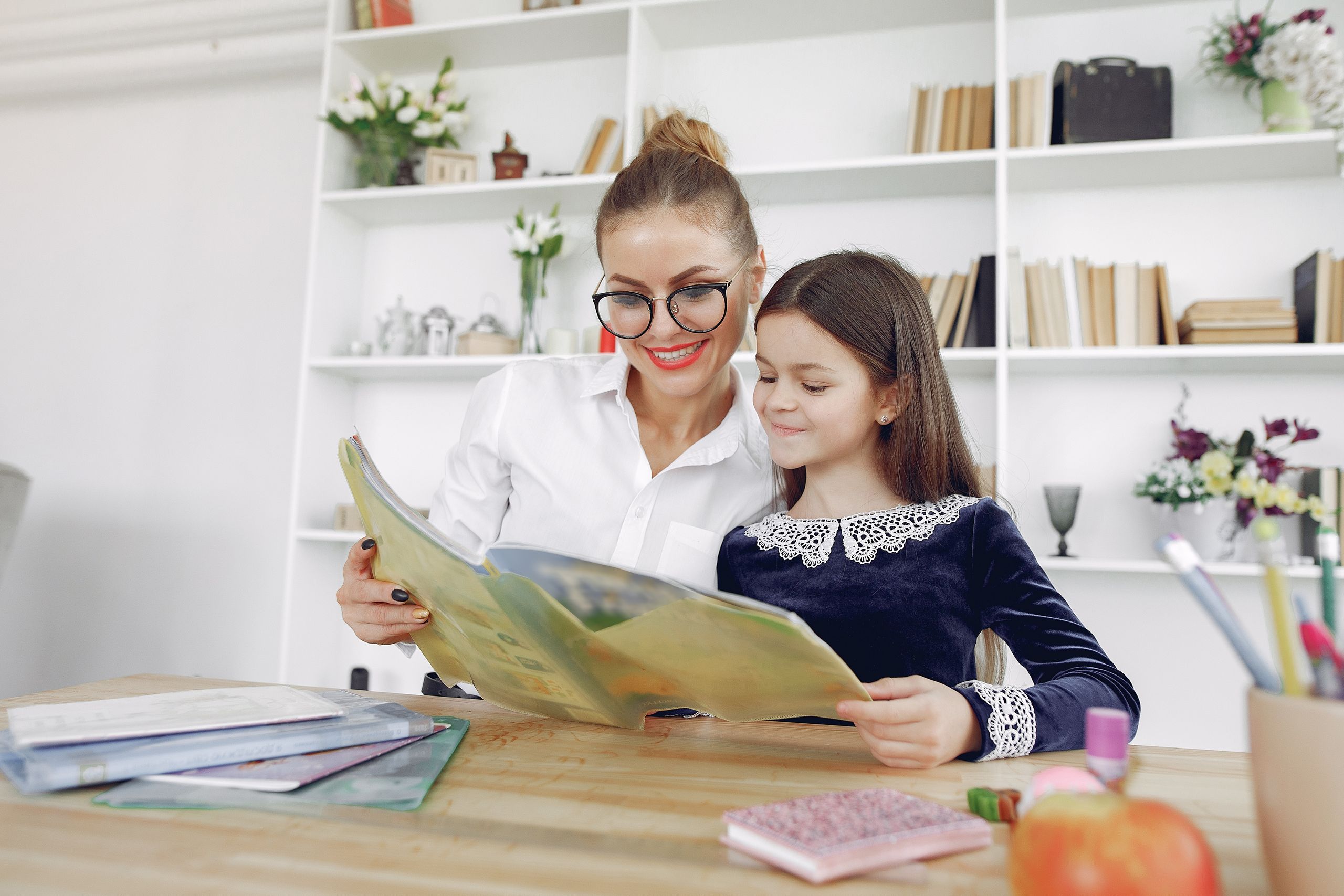 Smiling young woman in eyeglasses and girl in school uniform sitting at table and reading textbook together during preparation for classes in modern classroom