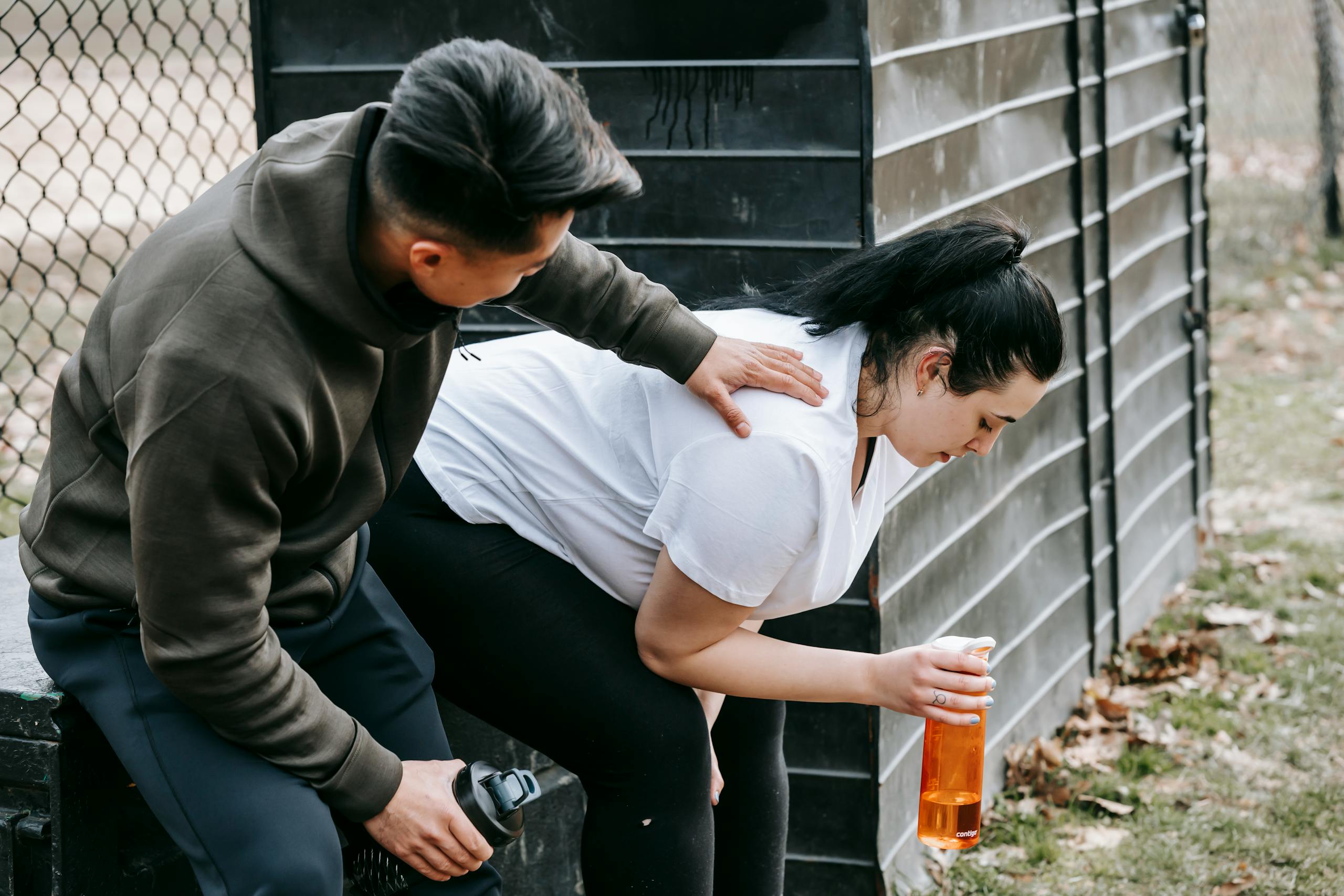 Sportsman clapping shoulder of plump tired female trainee sitting near net fence with bottle of water after training in autumn park
