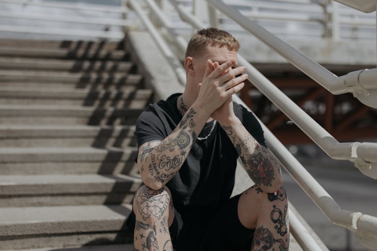 Stylish tattooed man sitting pensively on outdoor concrete stairs with hands clasped.