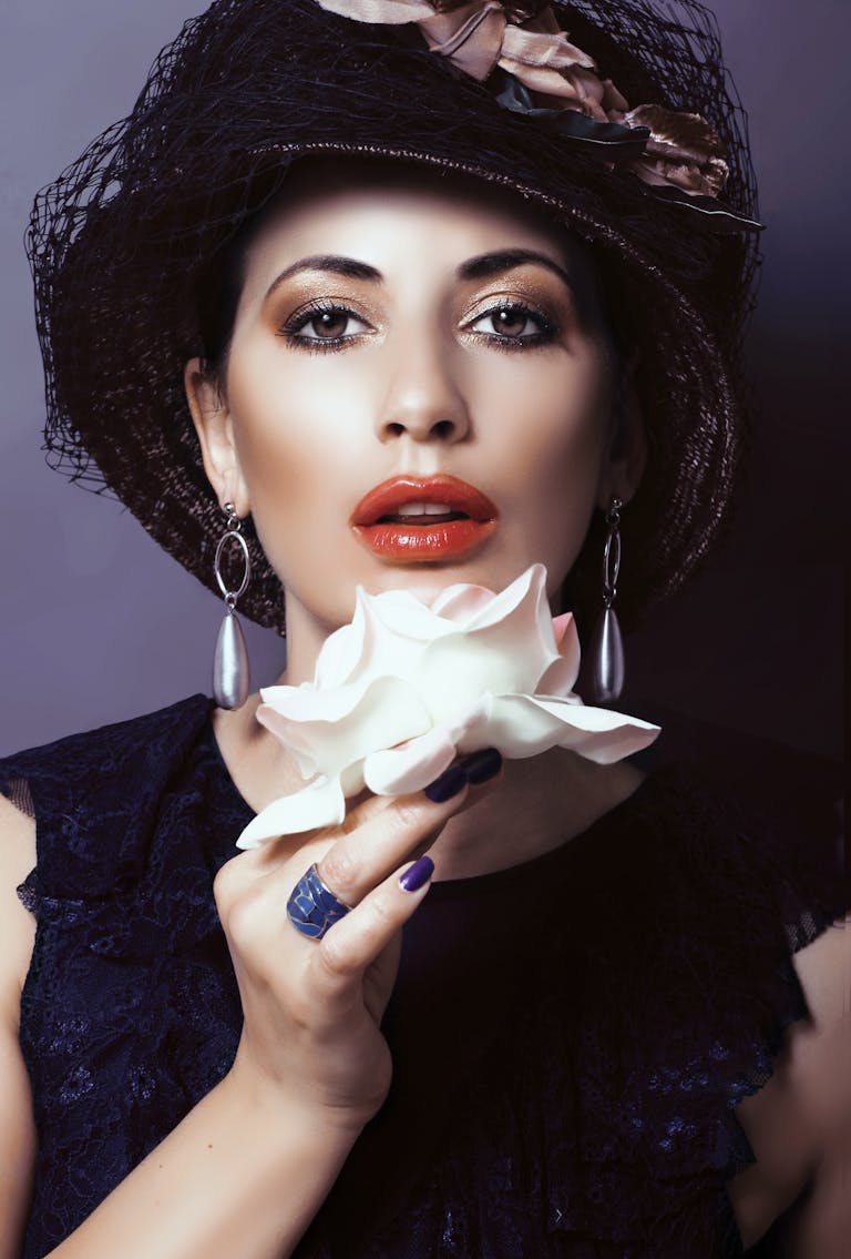 Stylish woman in a hat poses with a white rose, showcasing fashion and elegance in a studio setting.