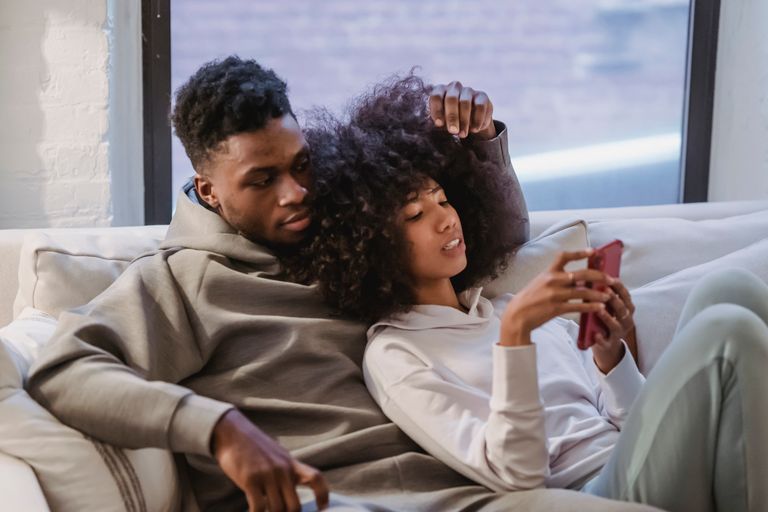 Stylish young African American lady with dark curly hair in casual outfit cuddling with boyfriend and messaging on smartphone while relaxing together on sofa at home