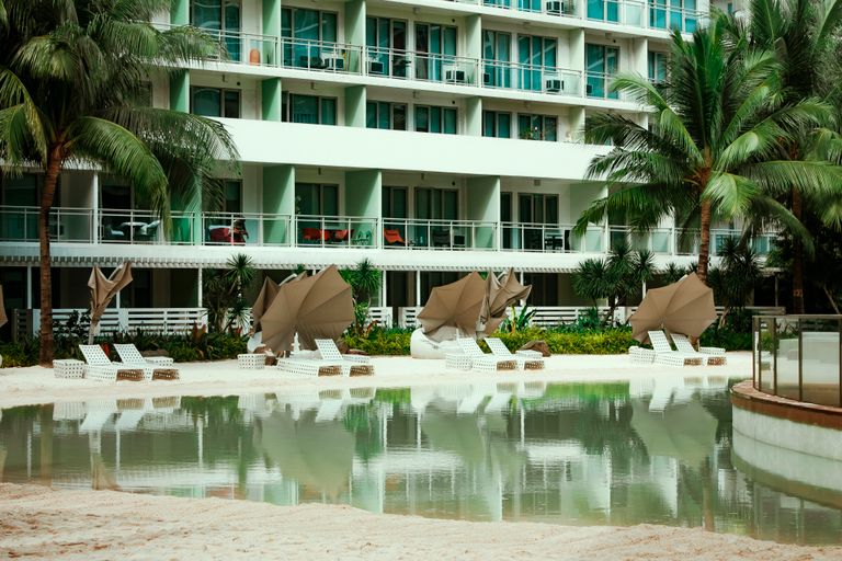 Sunbeds with umbrellas placed on sandy shore of swimming pool in yard of modern resort hotel with tropical trees