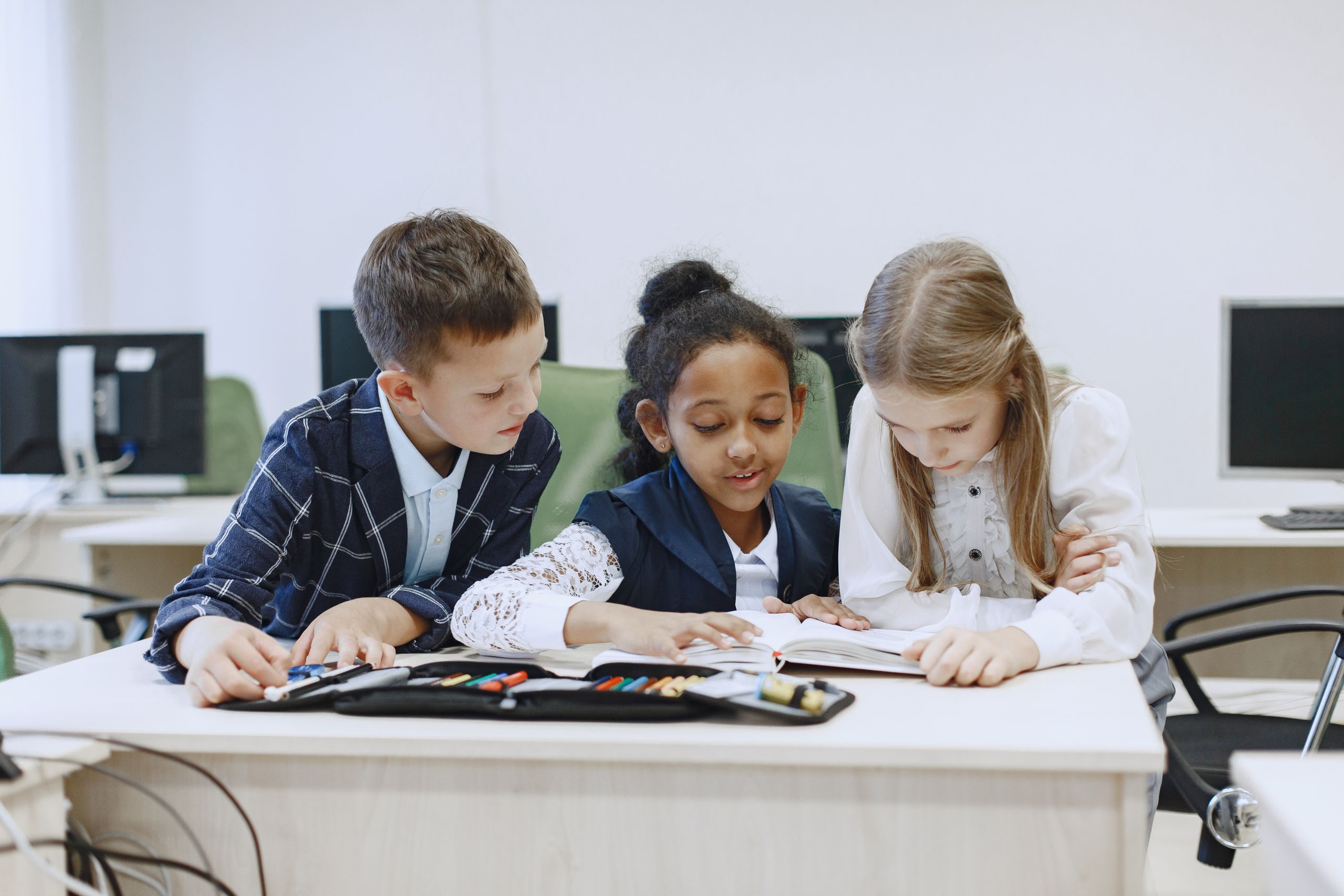 Three children working together at a desk in a classroom, focusing on a book.