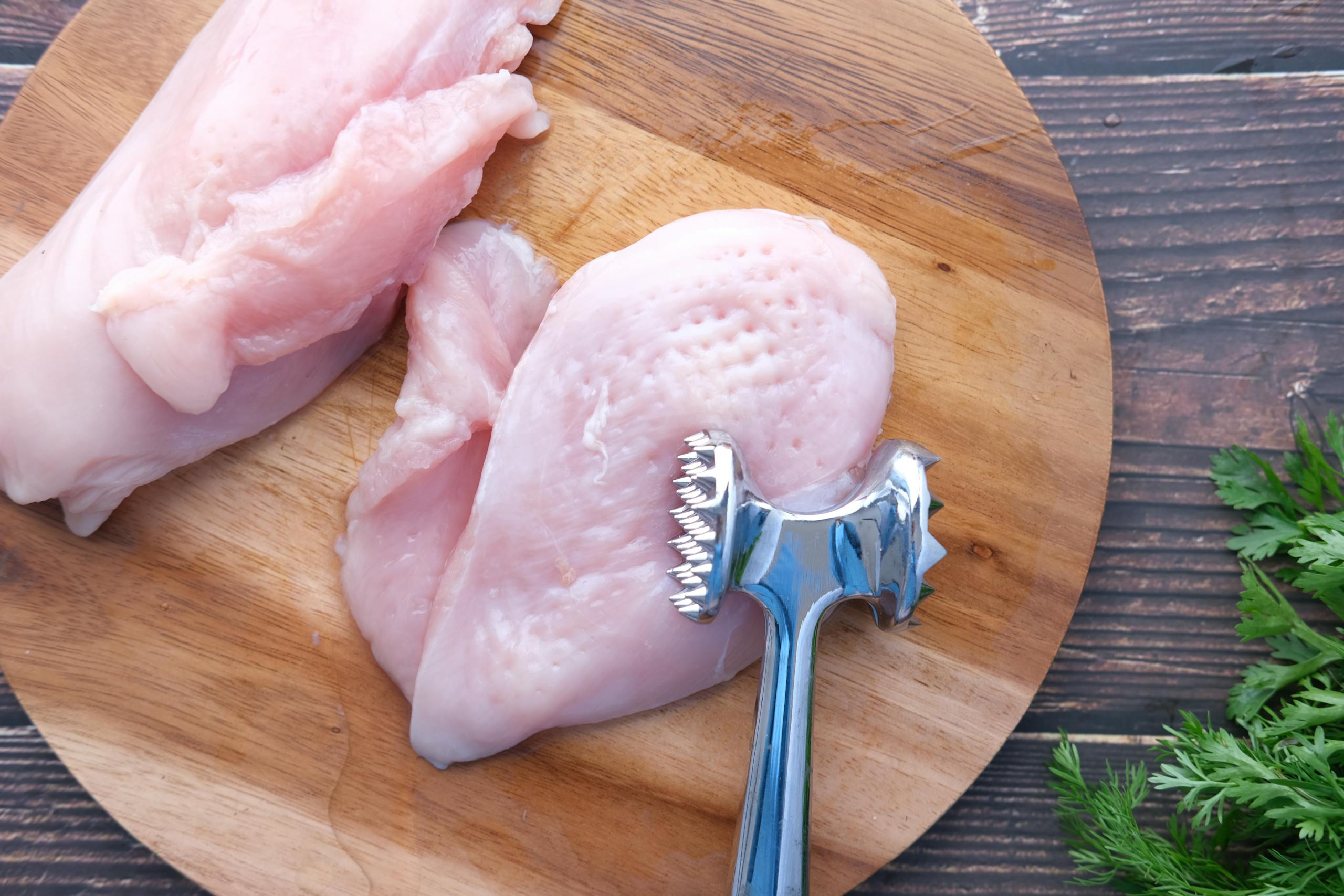Top view of raw chicken breasts on a wooden board with a meat mallet for tenderizing.