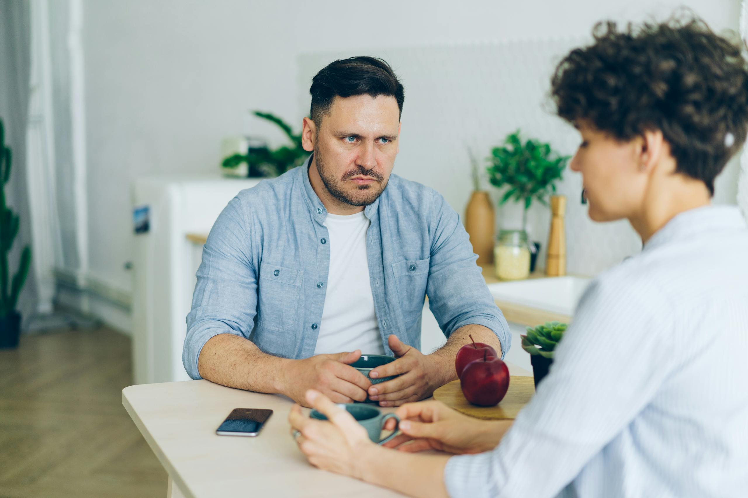 Two adults having a serious conversation at the dining table in a modern, well-lit kitchen.