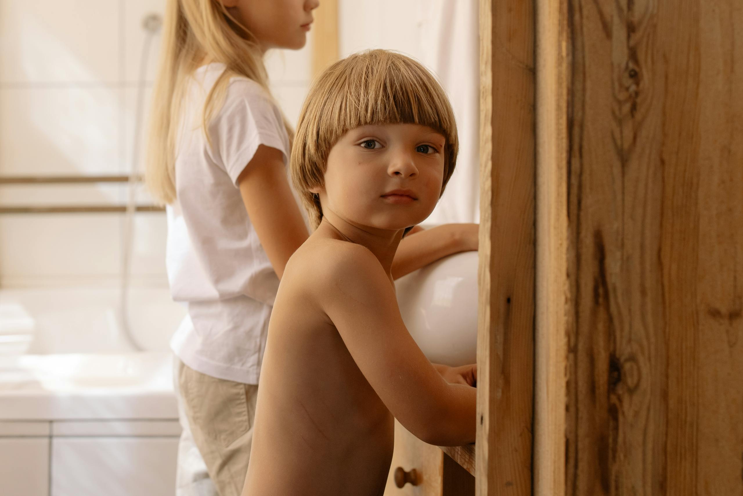 Two children in a bathroom; a young boy with a curious look and a girl facing away.