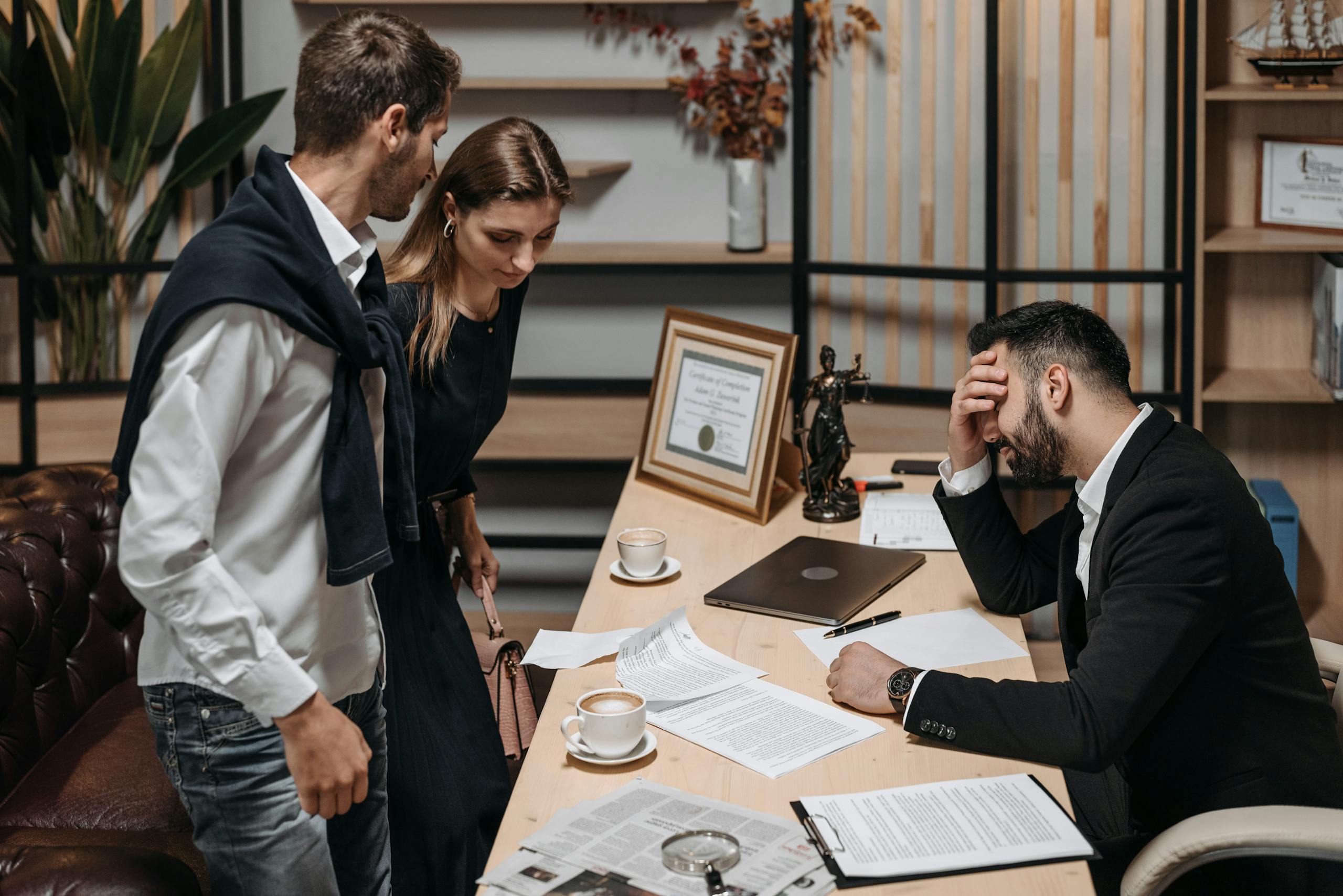 Two colleagues discuss reports with a stressed professional holding his head at an office desk.