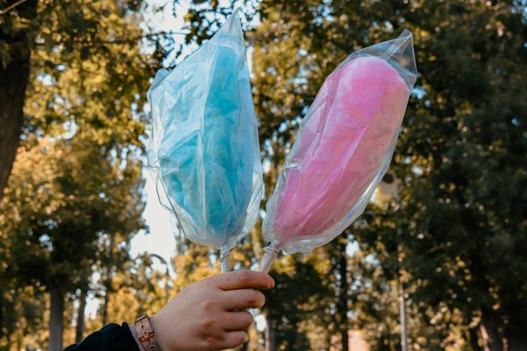 Two cotton candy sticks, one blue and one pink, held outdoors in a park setting.