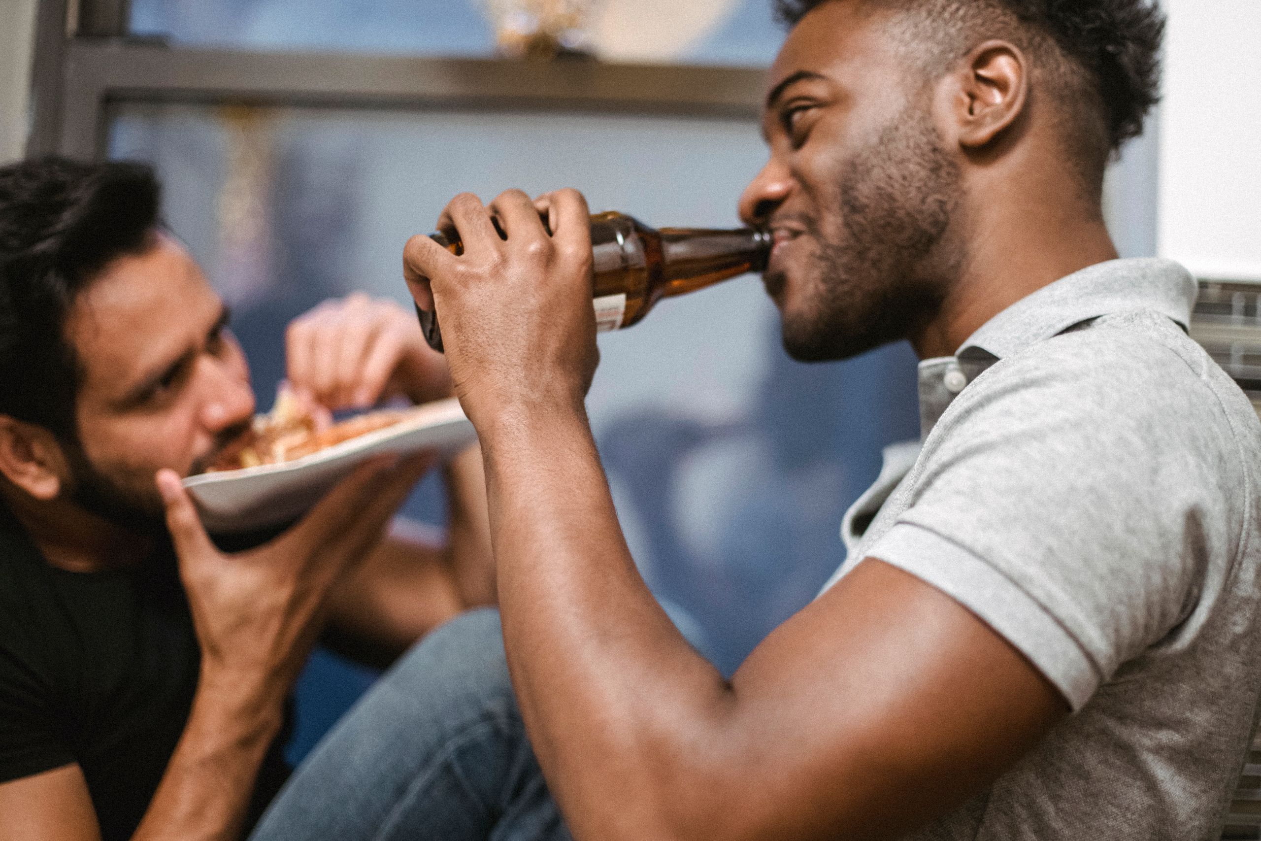 Two friends having a relaxed time, drinking beer and eating pizza in a cozy indoor setting.