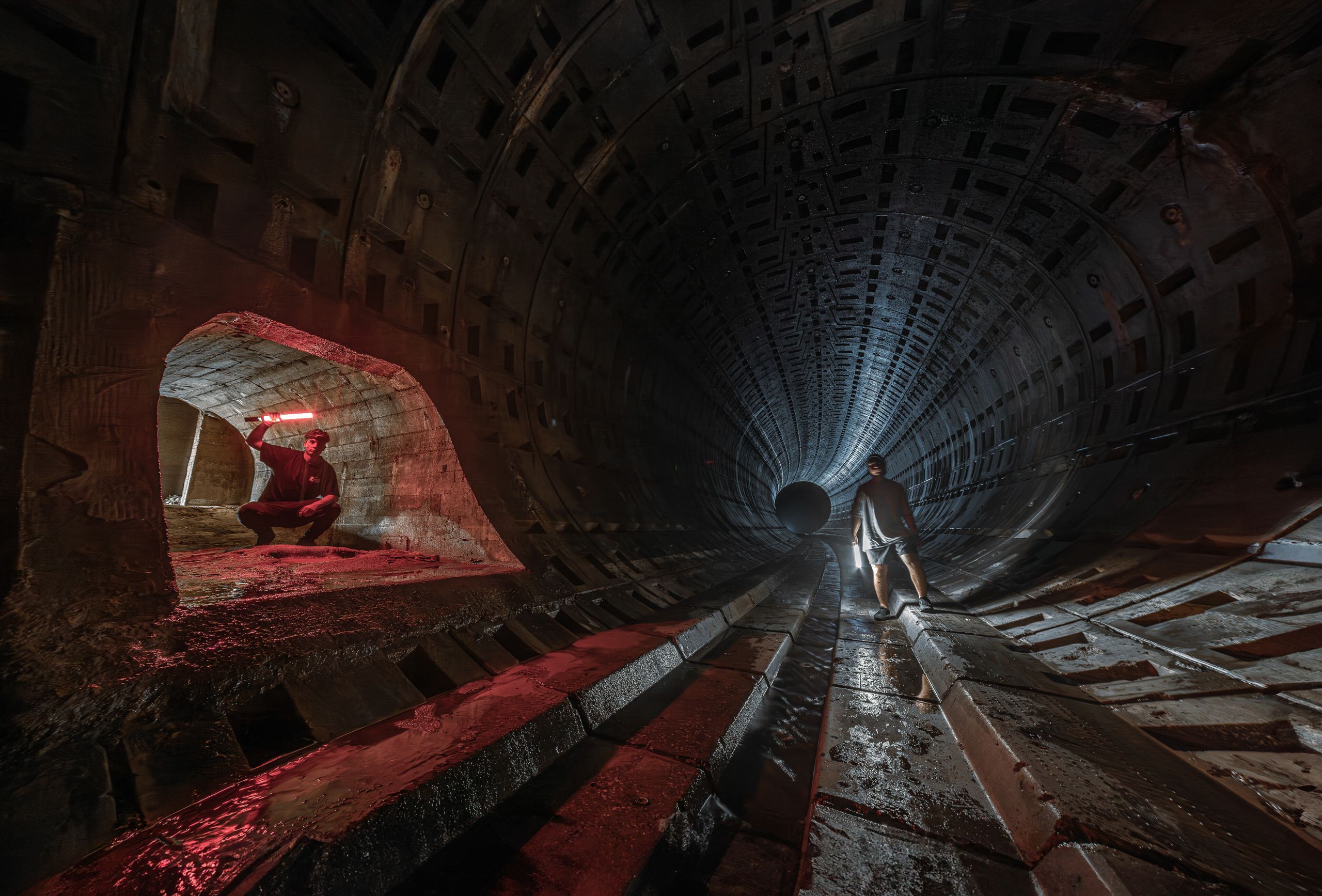 Two people explore a dimly lit underground tunnel with red lights, adding a mysterious and adventurous feel.