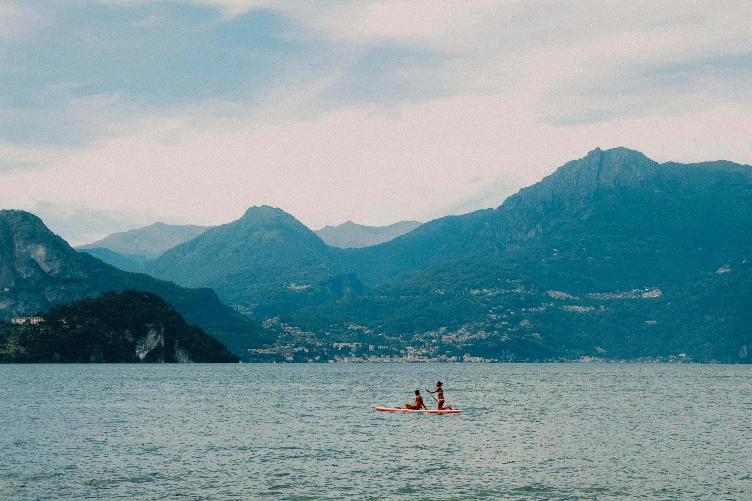 Two people kayaking on a tranquil lake surrounded by scenic mountains under a cloudy sky.