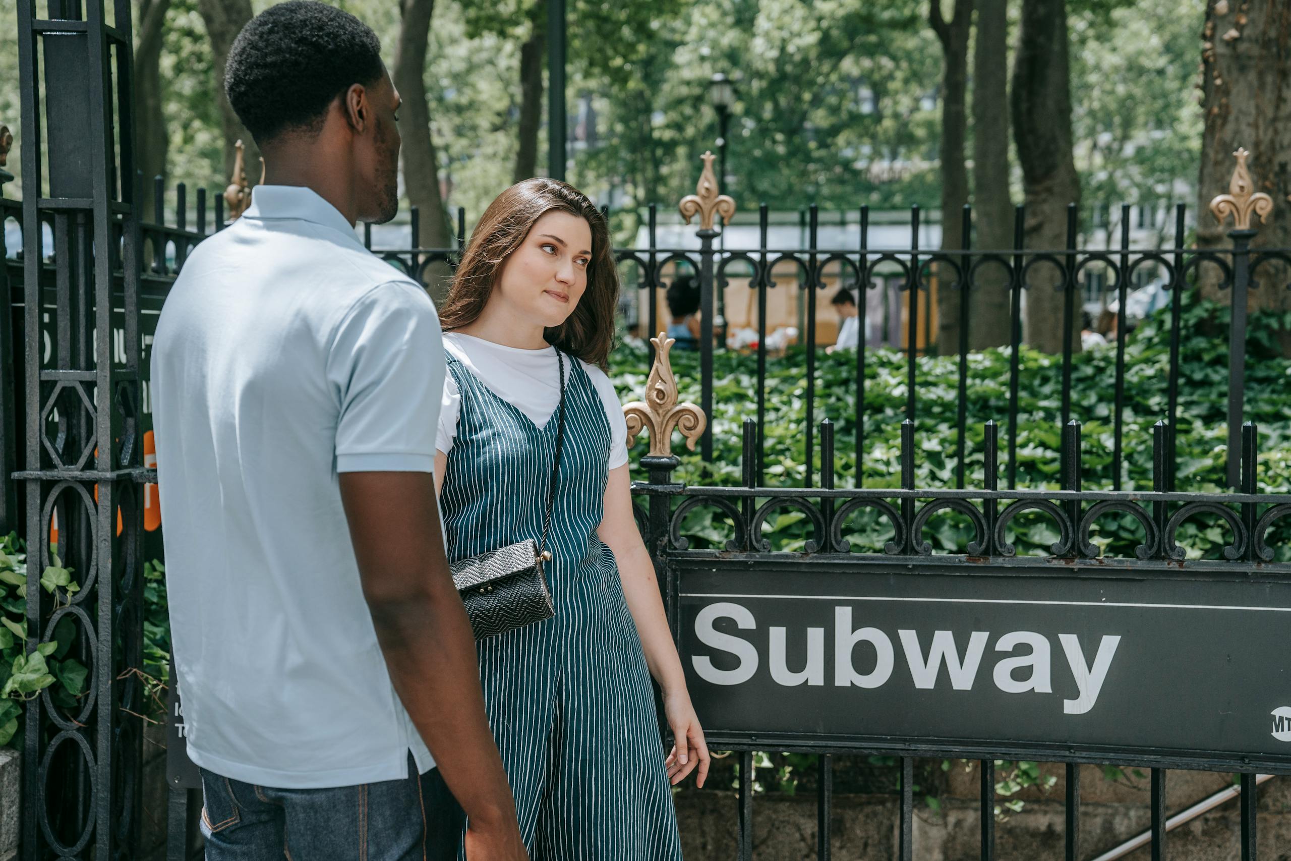 Two people standing outside a subway entrance on a sunny day, surrounded by greenery.