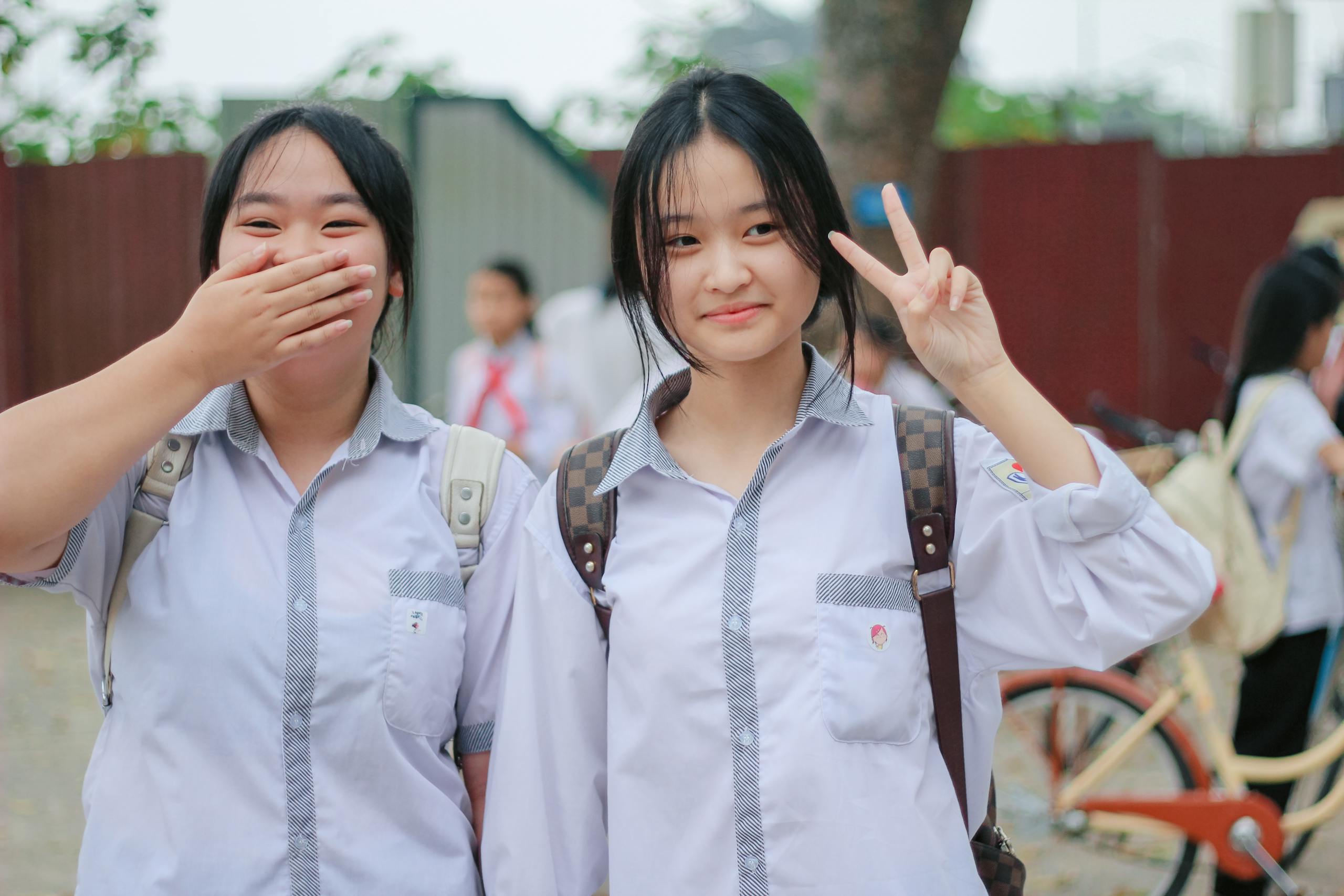 Two smiling school girls in uniforms outdoors, one gesturing peace.
