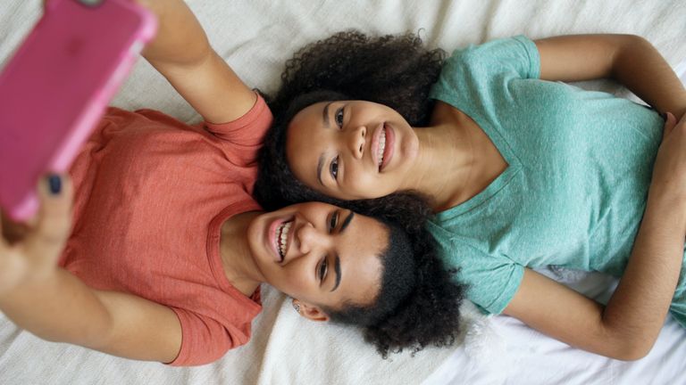 Two teenage girls lying on a bed, smiling while taking a selfie, capturing a joyful moment.