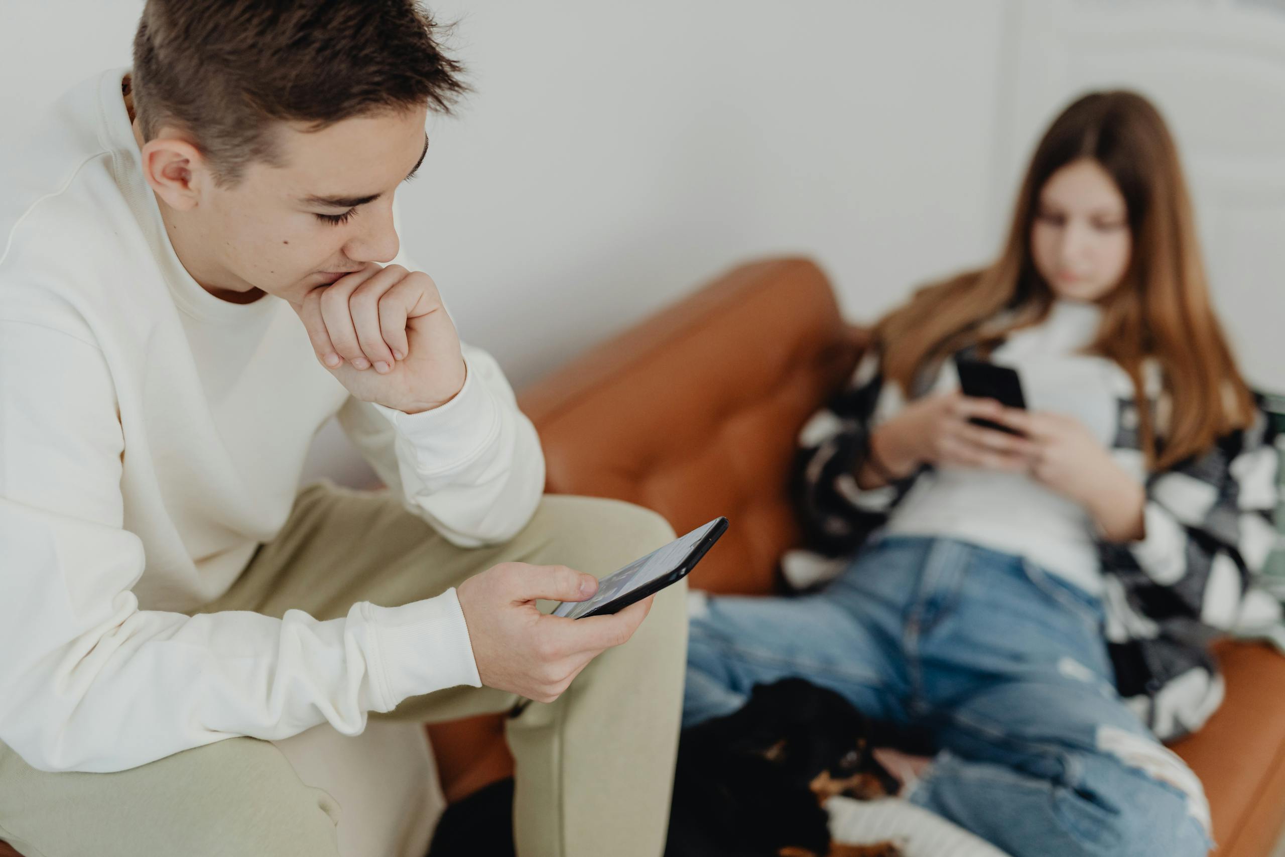 Two teenagers sitting casually on a couch, absorbed in their smartphones indoors.
