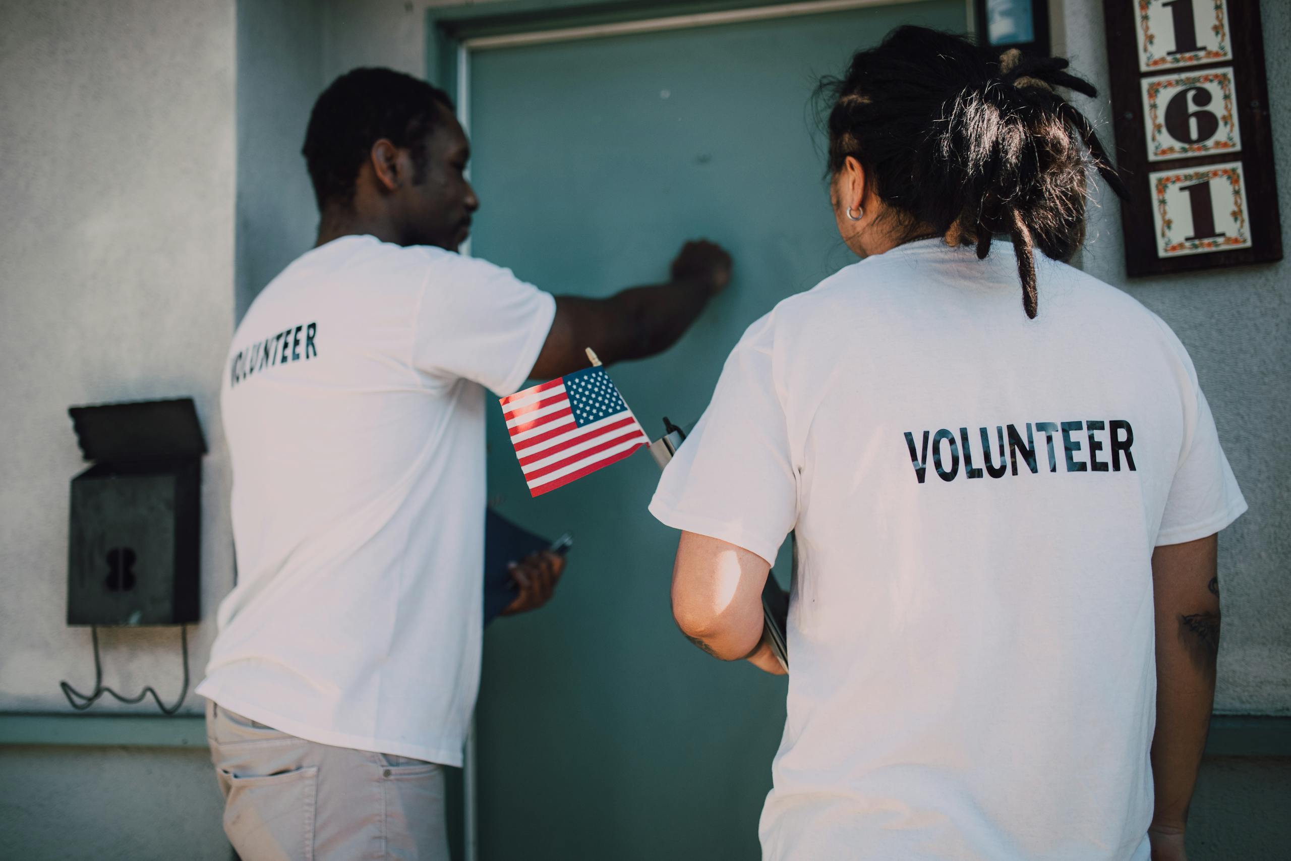 Two volunteers knocking on door during a community outreach event, holding a small American flag.