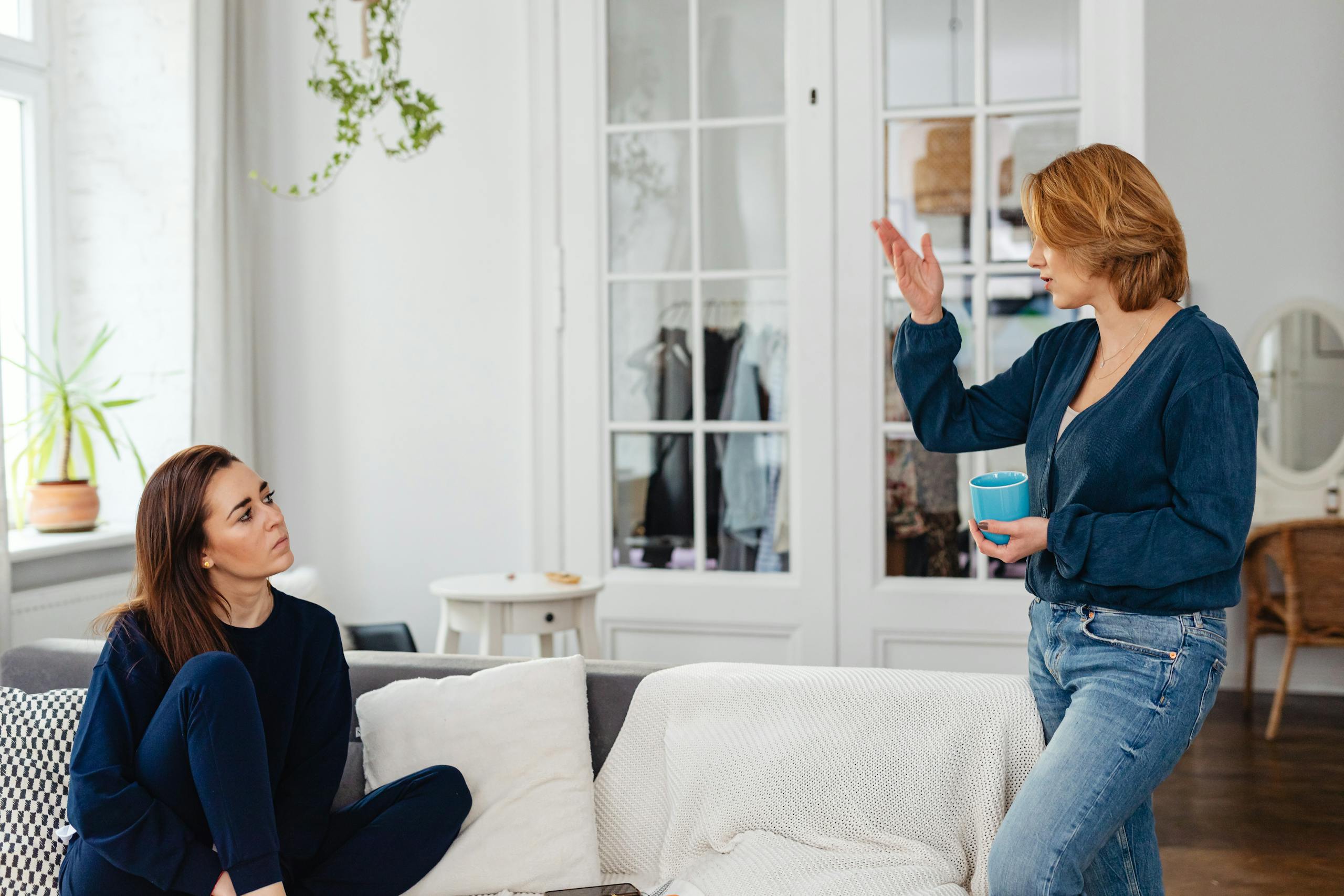 Two women engage in a friendly chat in a stylish living room, one sitting and one standing with a mug.