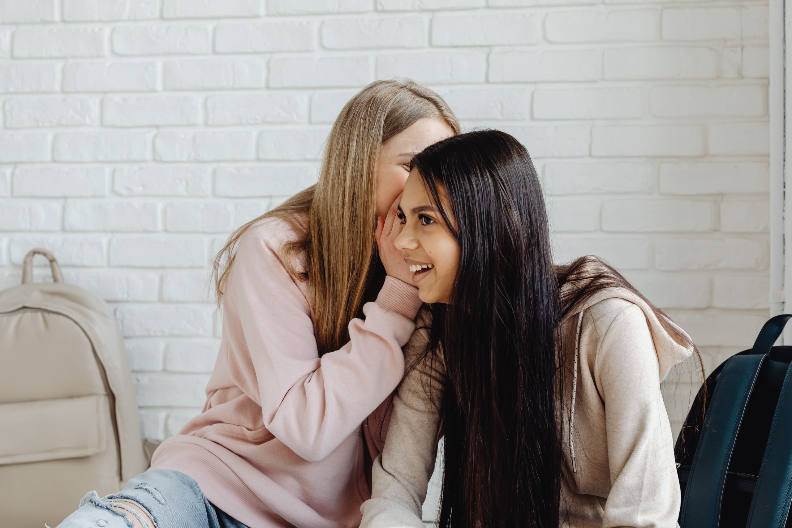 Two women in casual attire whisper and laugh against a white brick wall.