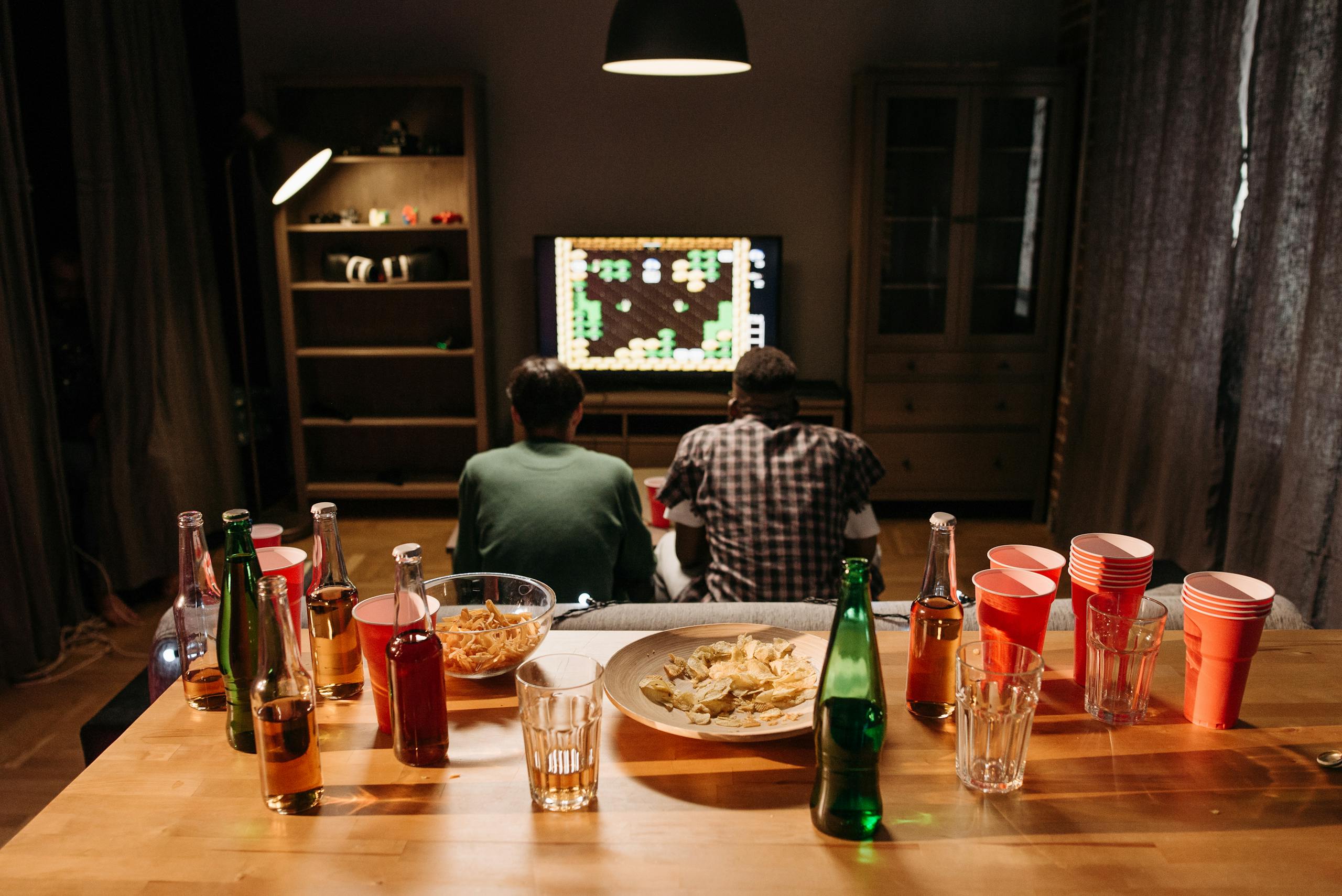 Two young men play video games in a cozy living room with snacks and drinks on the table.